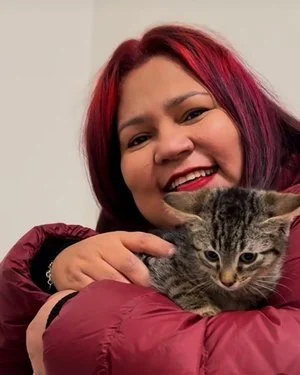 Una mujer con cabello de color rojo y violeta abrazando a un gato gris con rayas.