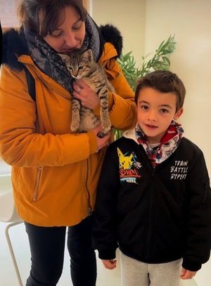 Niña sosteniendo un gato y un niño sonriendo al lado, en un ambiente interior.