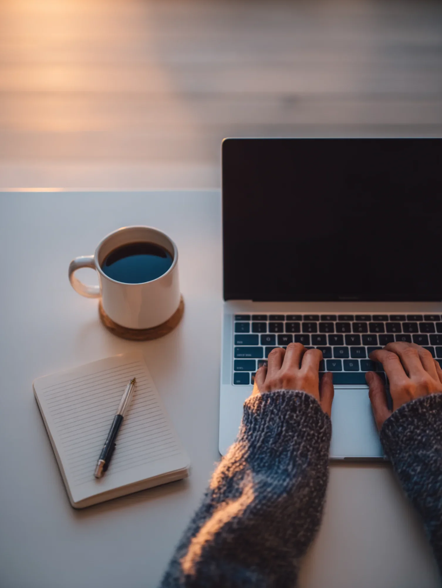Person working on a laptop with a cup of coffee, a notebook, and a pen on a white desk at sunrise or sunset.