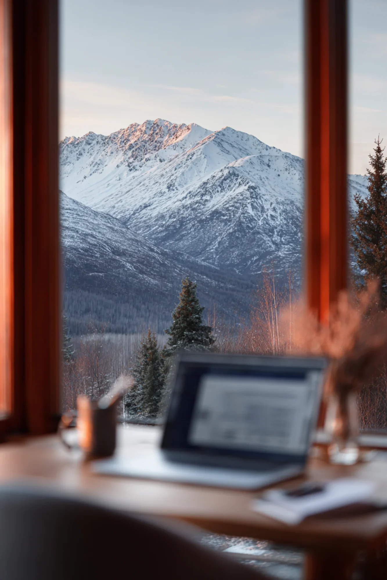 A window view of snow-covered mountains with trees in the foreground, viewed from a cozy indoor space with a blurred laptop and desk in the foreground.
