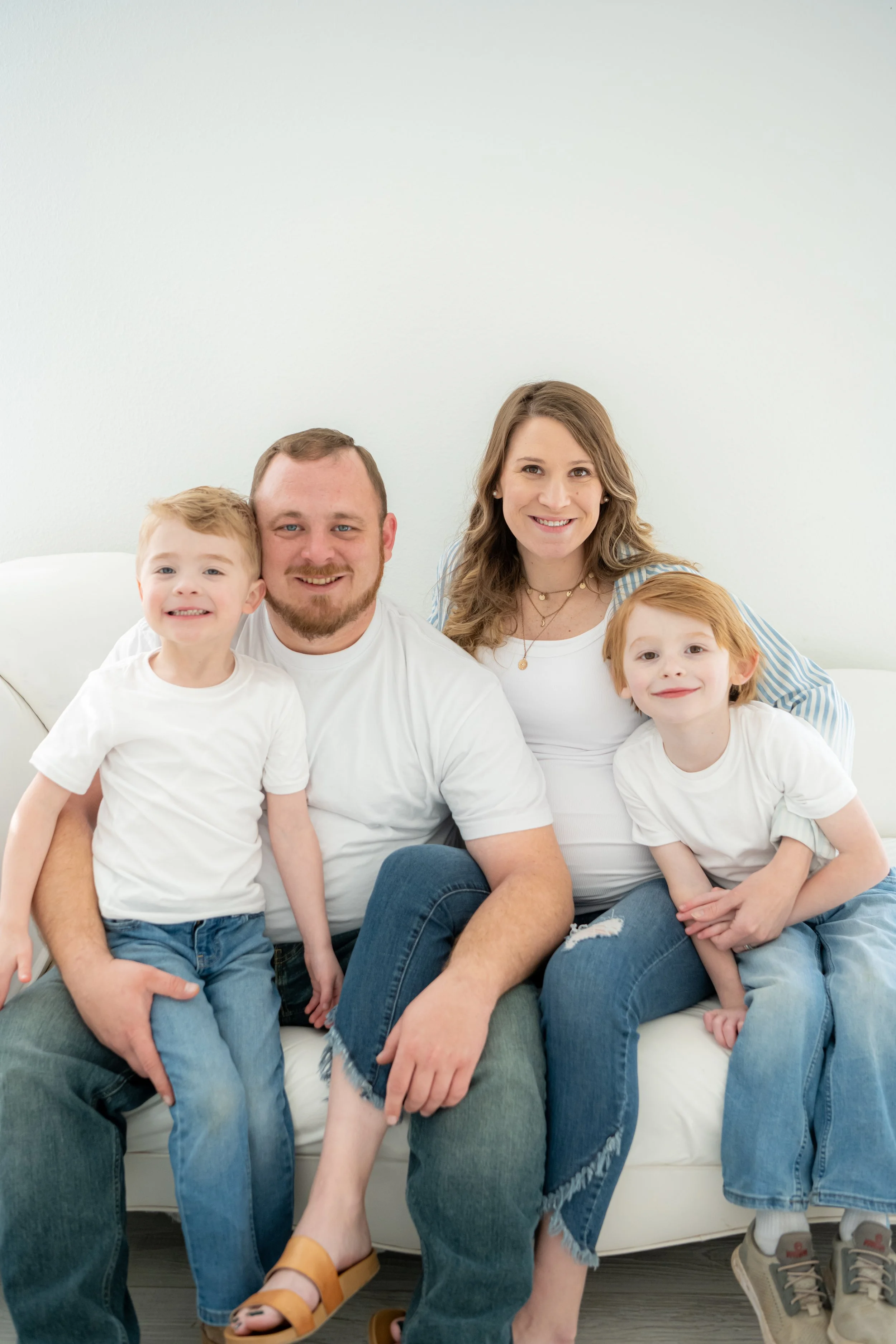 A smiling family of four sitting together on a white couch, including a mother, father, and two young boys, all wearing casual white and blue clothing in a bright room.