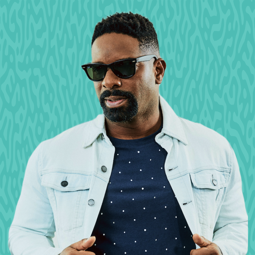 Man with dark hair, beard, and sunglasses wearing a light denim jacket over a navy T-shirt with white dots, standing against a teal background with abstract patterns.