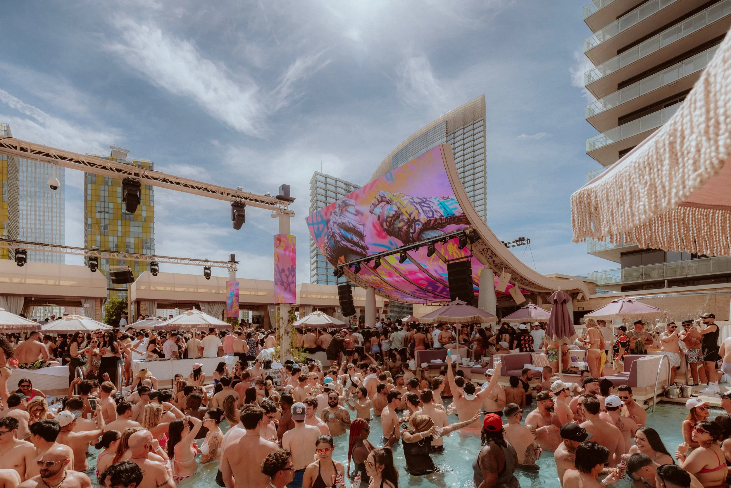 Crowd of people enjoying a pool party at an outdoor venue with a large stage and digital screens, surrounded by modern high-rise buildings.
