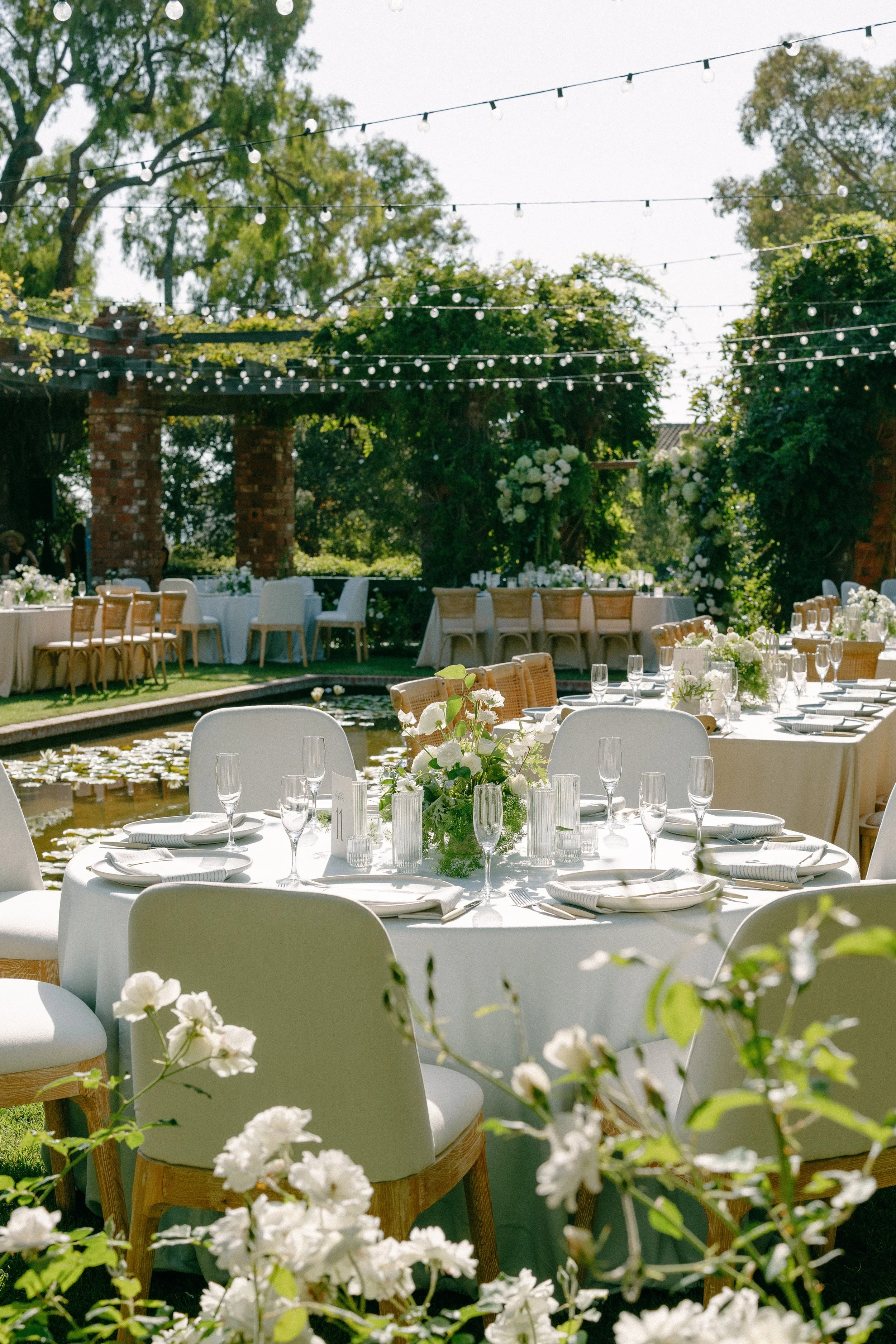Elegant outdoor wedding reception with round tables, white tablecloths, floral centerpieces, silverware, glassware, surrounded by chairs, in a lush garden with string lights overhead and trees in the background.