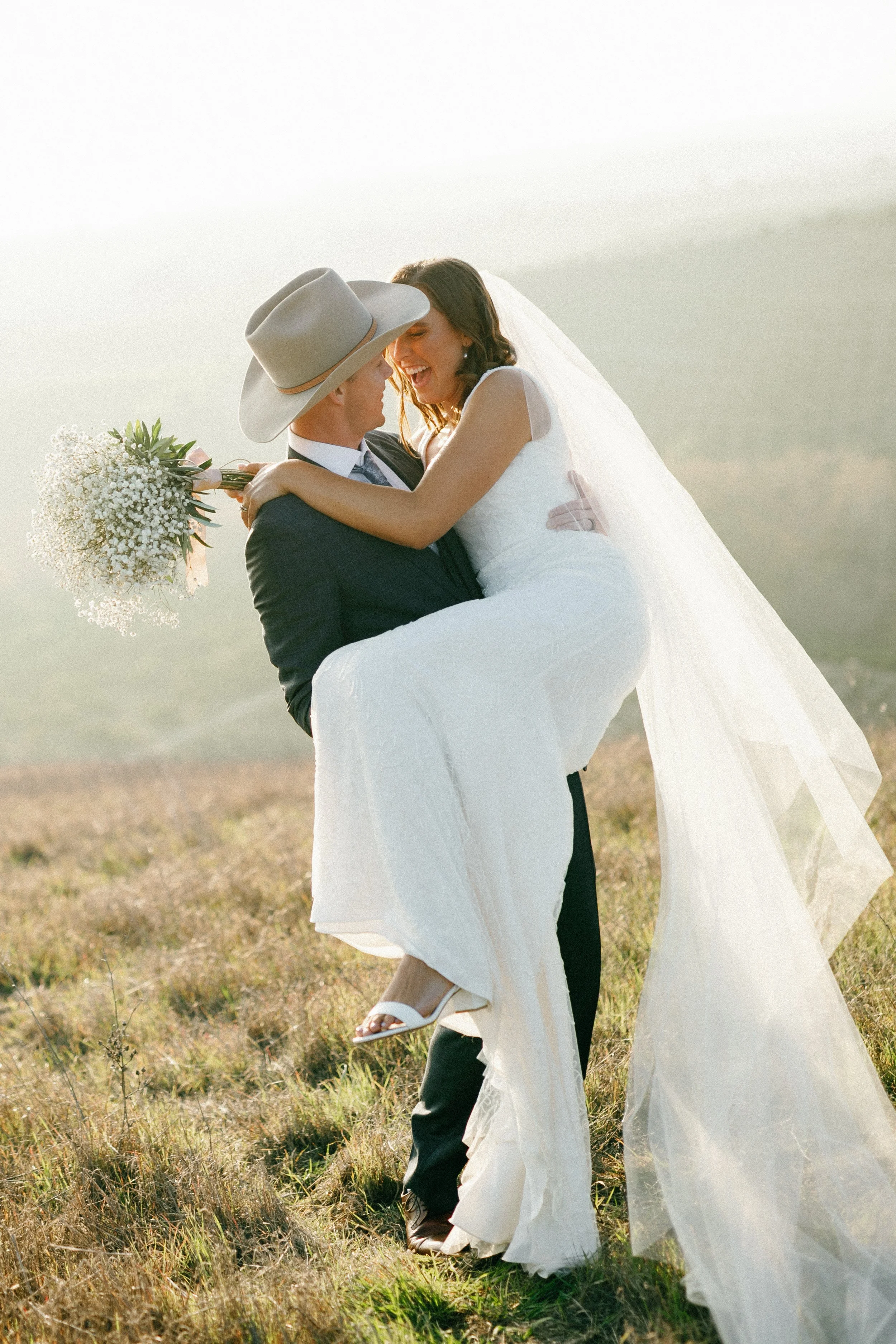 A man in a suit and cowboy hat carrying a woman in a wedding dress and veil, holding a bouquet, outdoor during sunset.