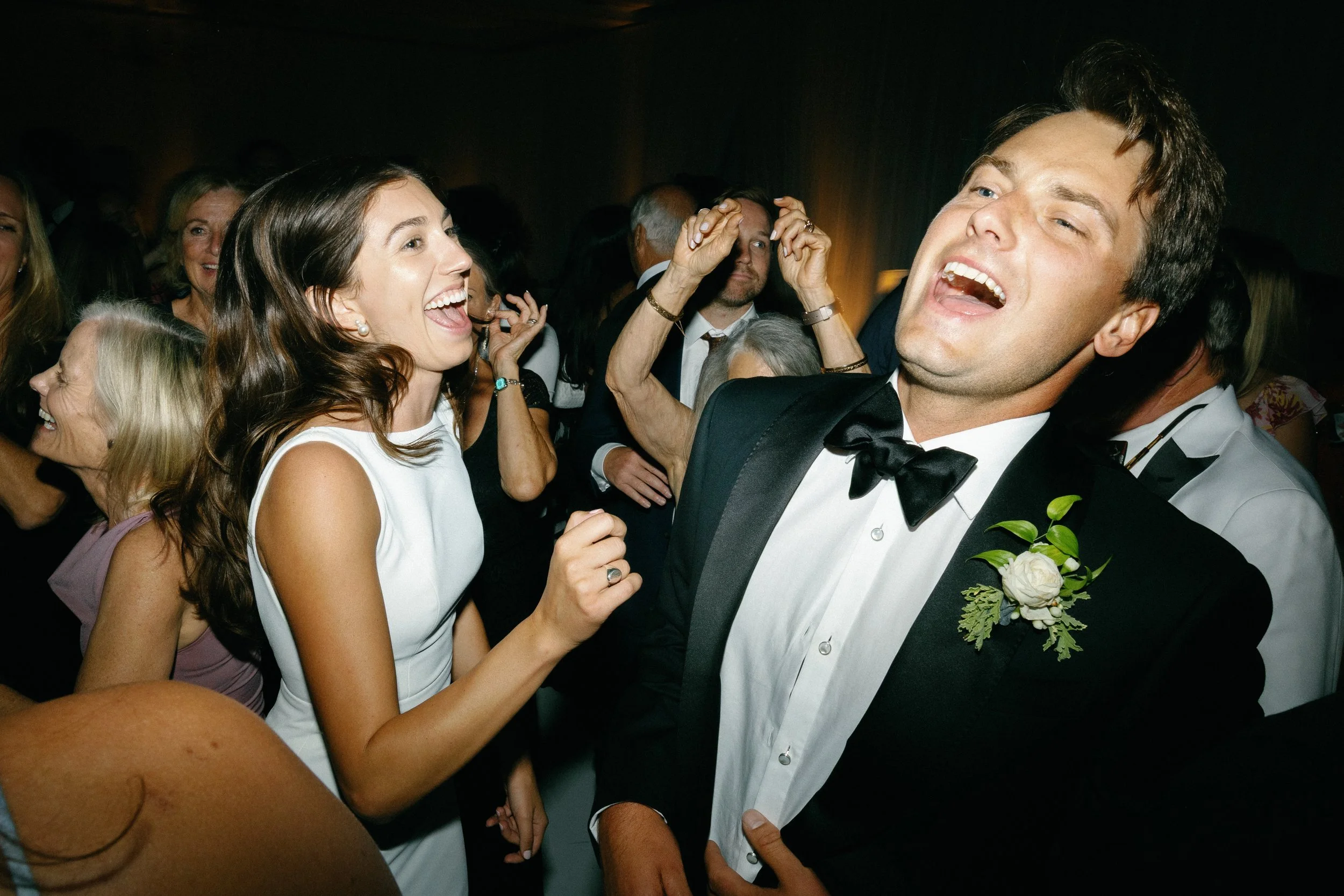 People dancing and laughing at a wedding or party, with a woman in a white dress and a man in a tuxedo in the foreground.