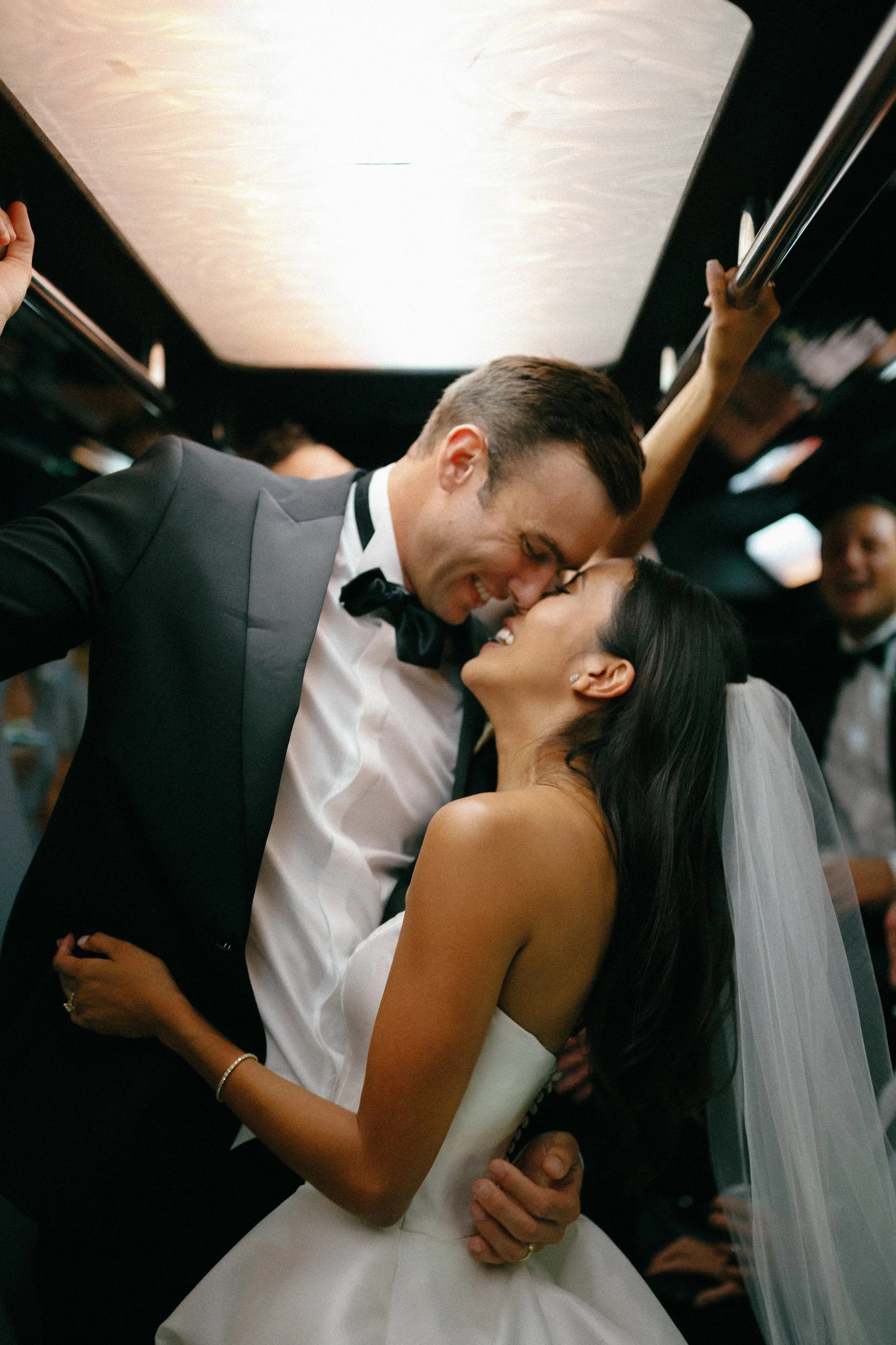 A newlywed couple sharing a romantic moment on a bus, with the groom leaning down to kiss the bride, who is smiling with her eyes closed.