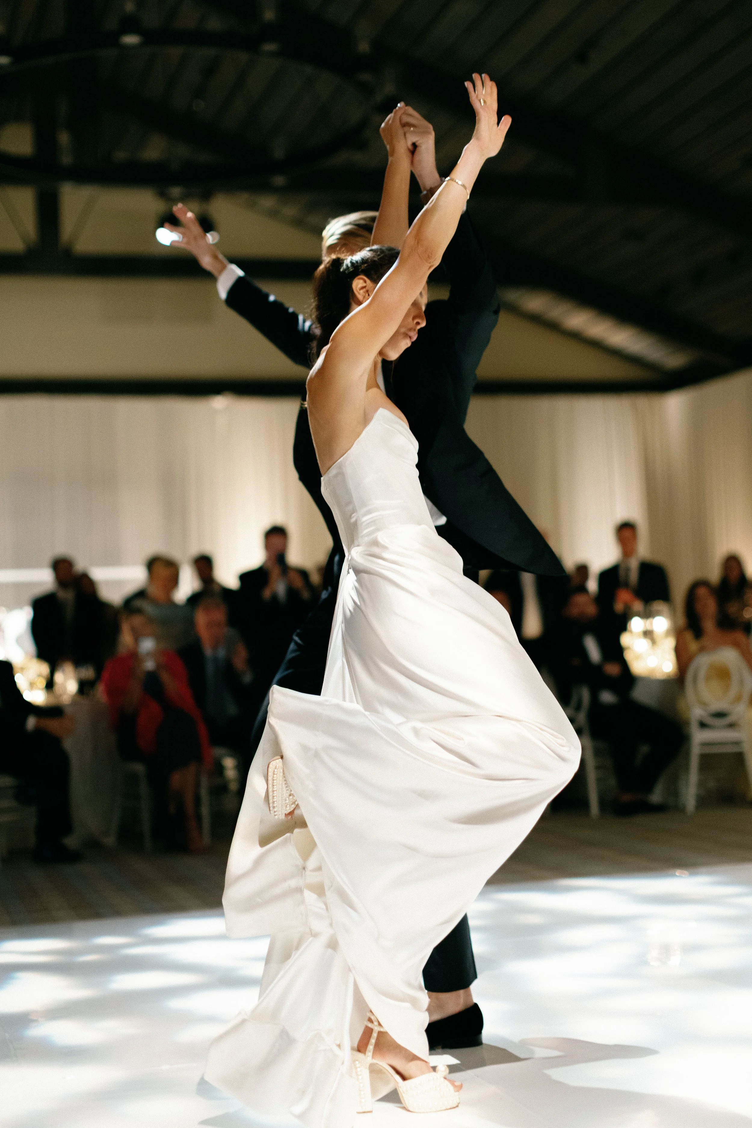 A bride in a white wedding dress and a groom in a black tuxedo are dancing closely, with their hands raised above their heads, on a dance floor at a wedding reception. Guests are seated at tables in the background, watching the dance.