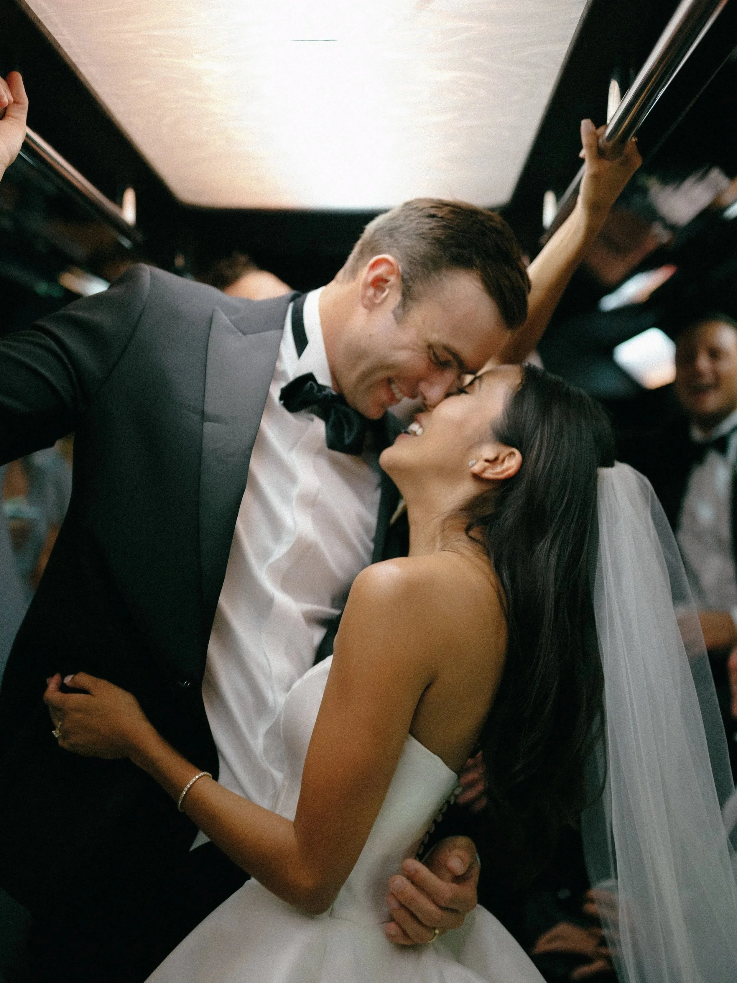 A newlywed couple sharing a romantic moment on a bus, with the groom leaning down to kiss the bride, who is smiling with her eyes closed.