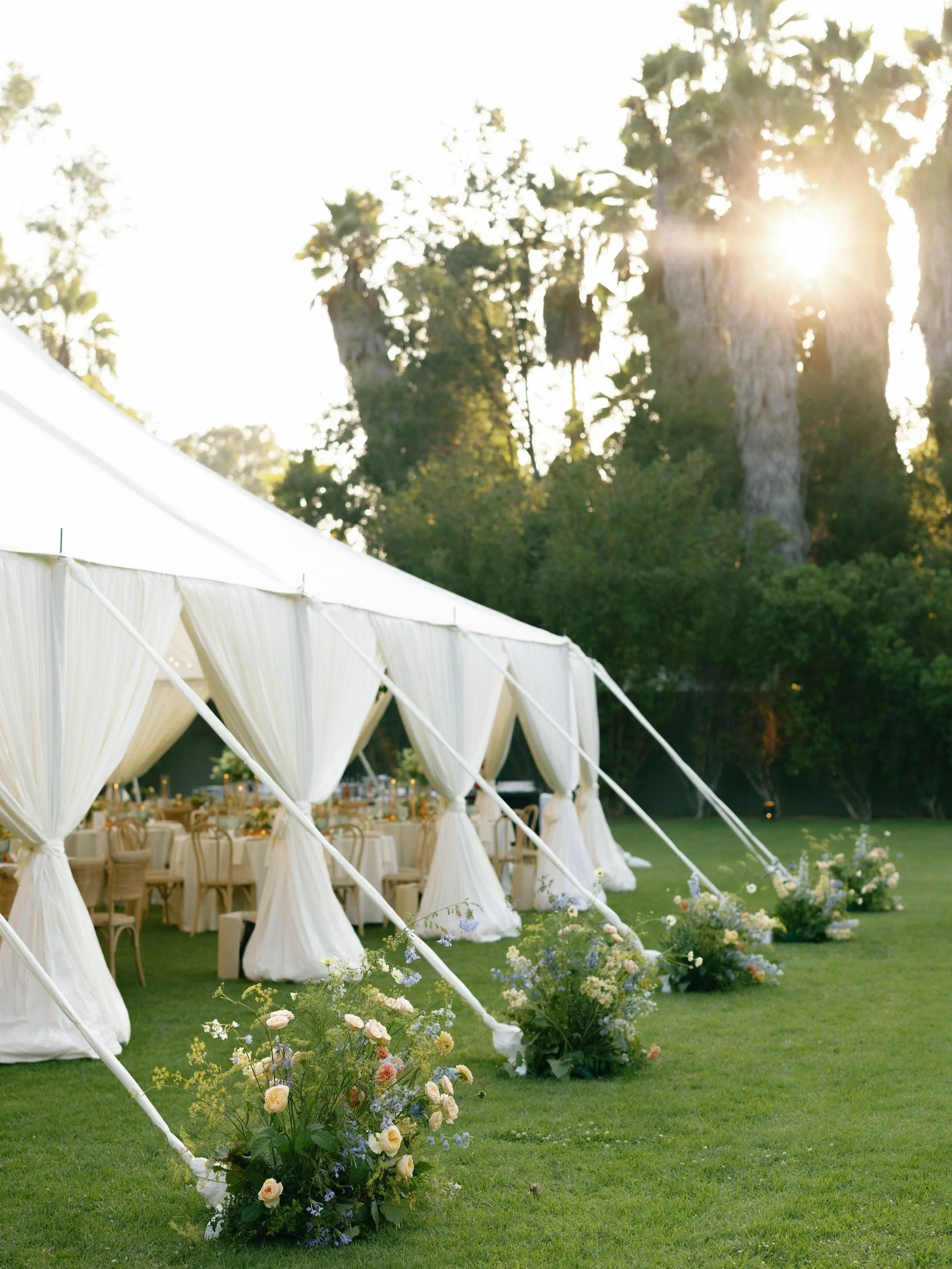 An outdoor event setup with a large white tent, decorated with floral arrangements at the entrance, on a green lawn with tall trees and sunlight shining through.