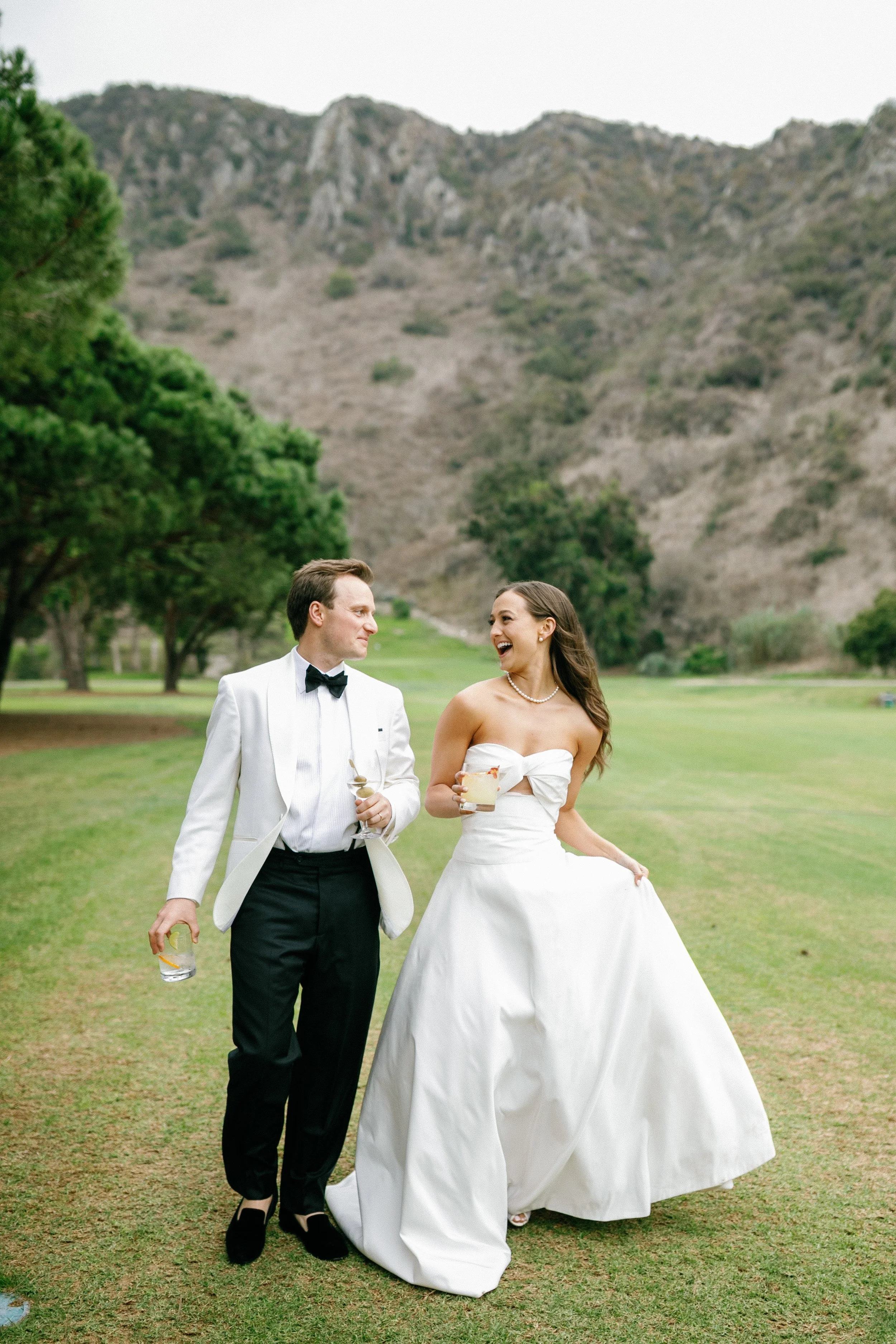 A bride and groom walking outdoors on a grassy area, smiling and holding drinks, with a mountainous background and trees.