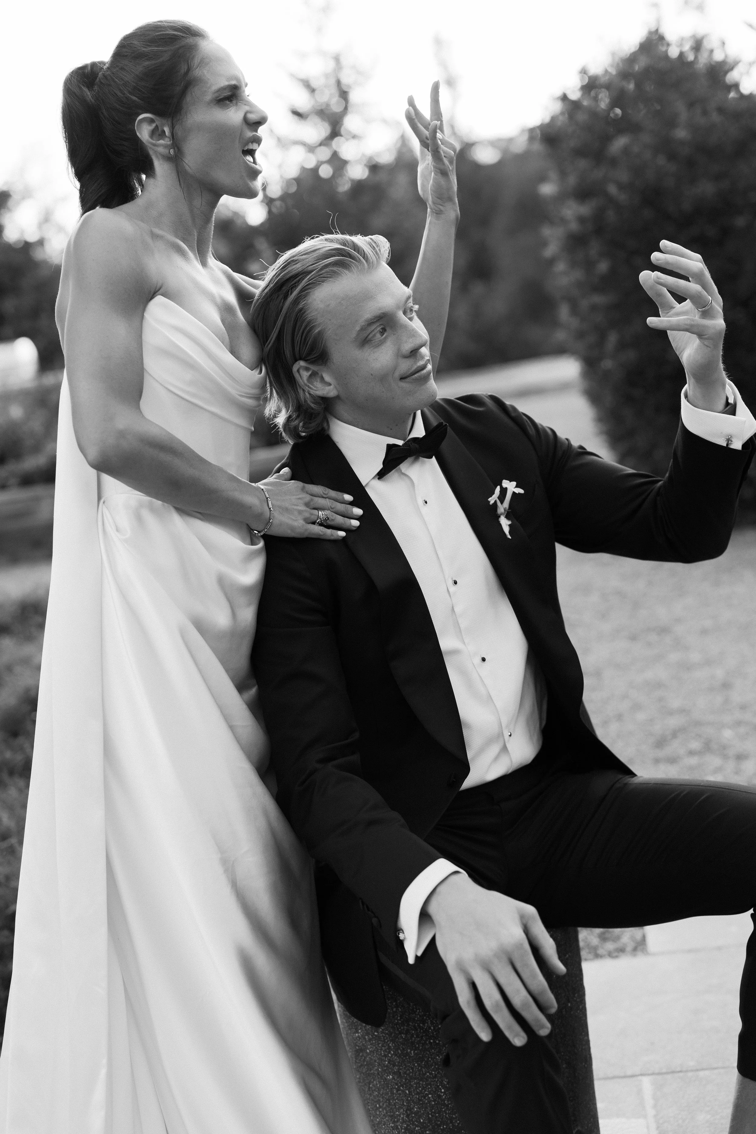 A black-and-white photo of a woman in an off-the-shoulder dress and a man in a tuxedo, outdoors. The woman appears to be yelling or expressing emotion, while the man is seated and taking a selfie or photo with his phone.