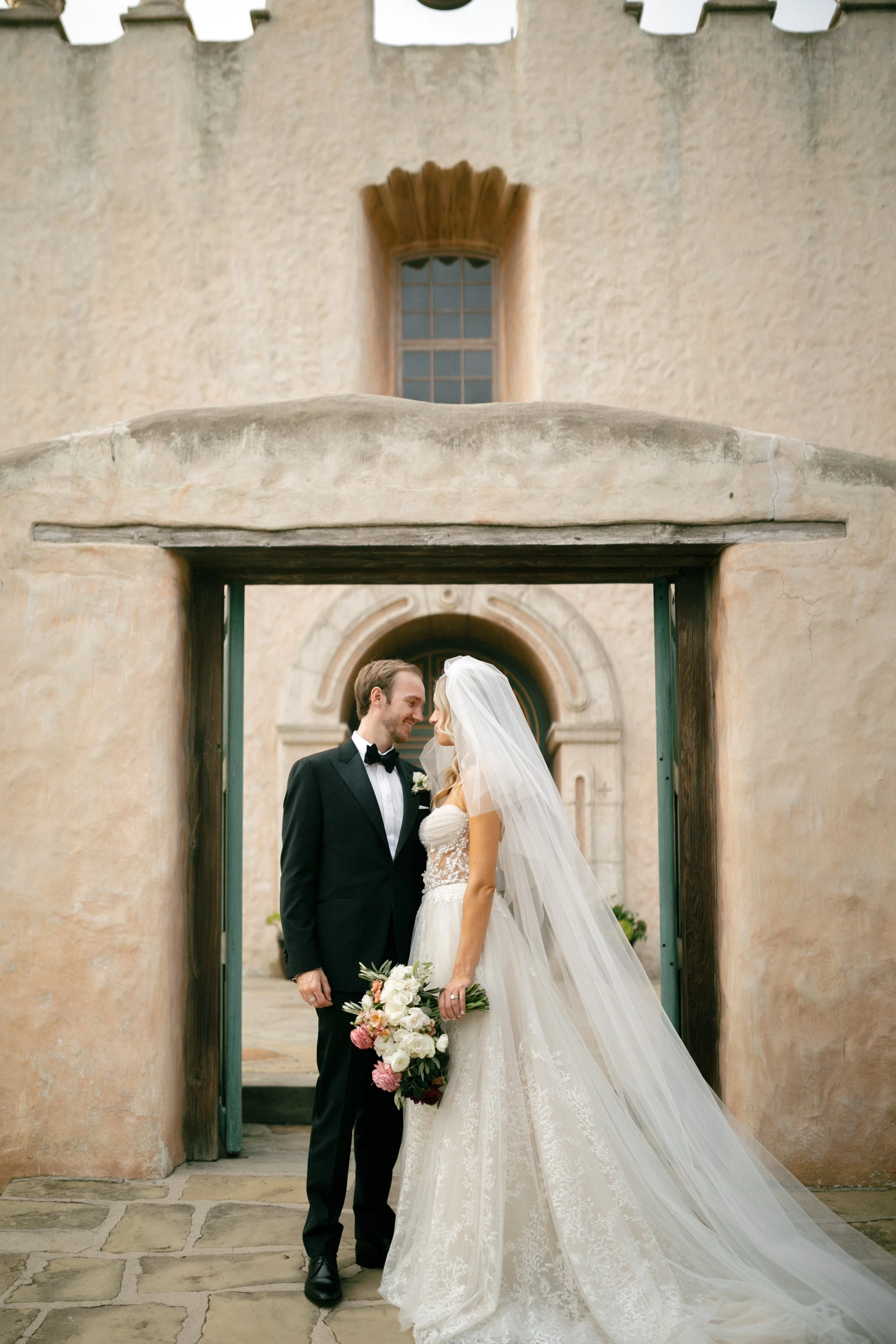 A bride and groom standing close together, smiling, outside a building with rustic walls and an arched doorway, the bride holding a bouquet of flowers.
