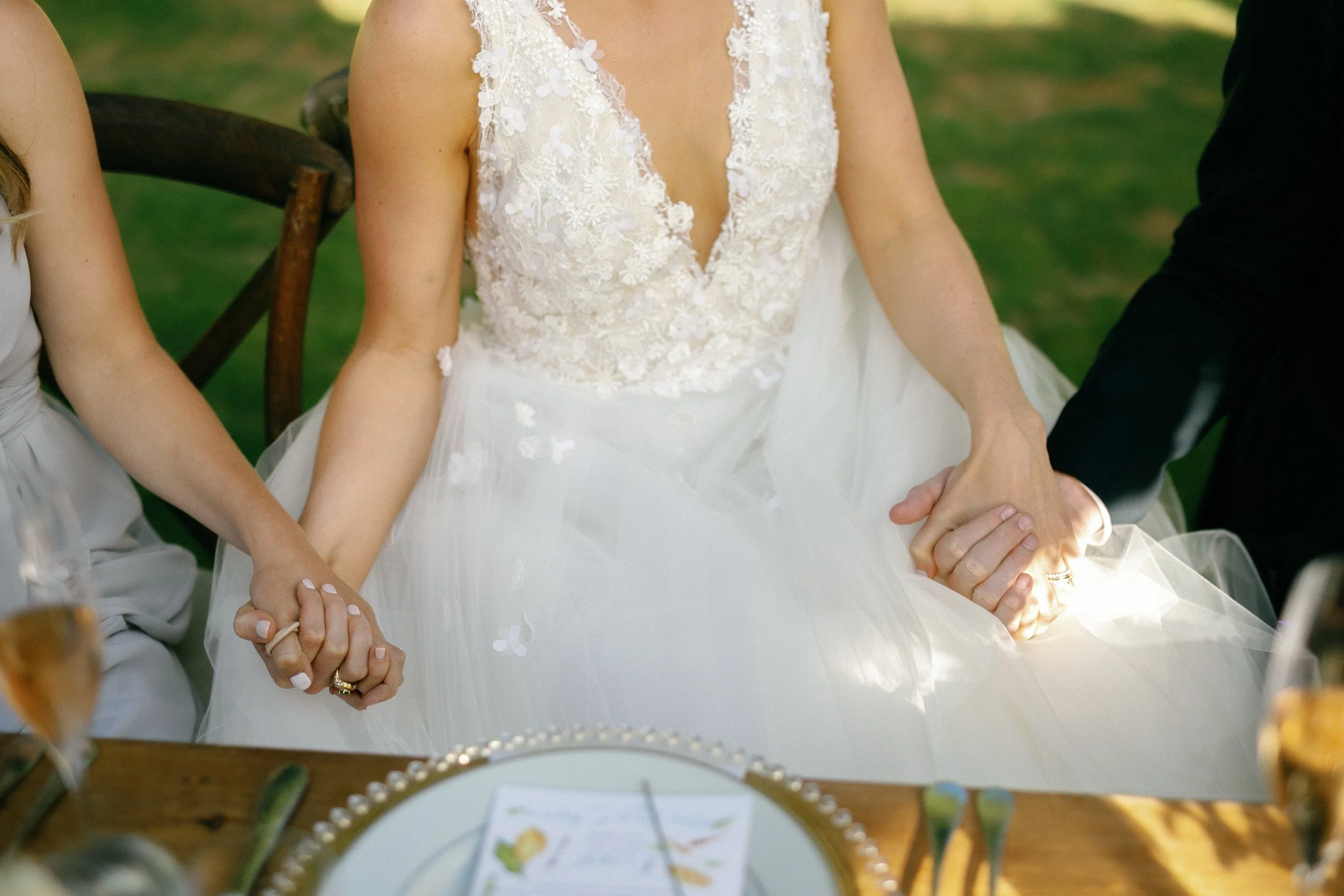 Wedding ceremony with bride holding hands with another person, wearing a white lace wedding dress, seated at a table with a plate and glasses, outdoors with green background.