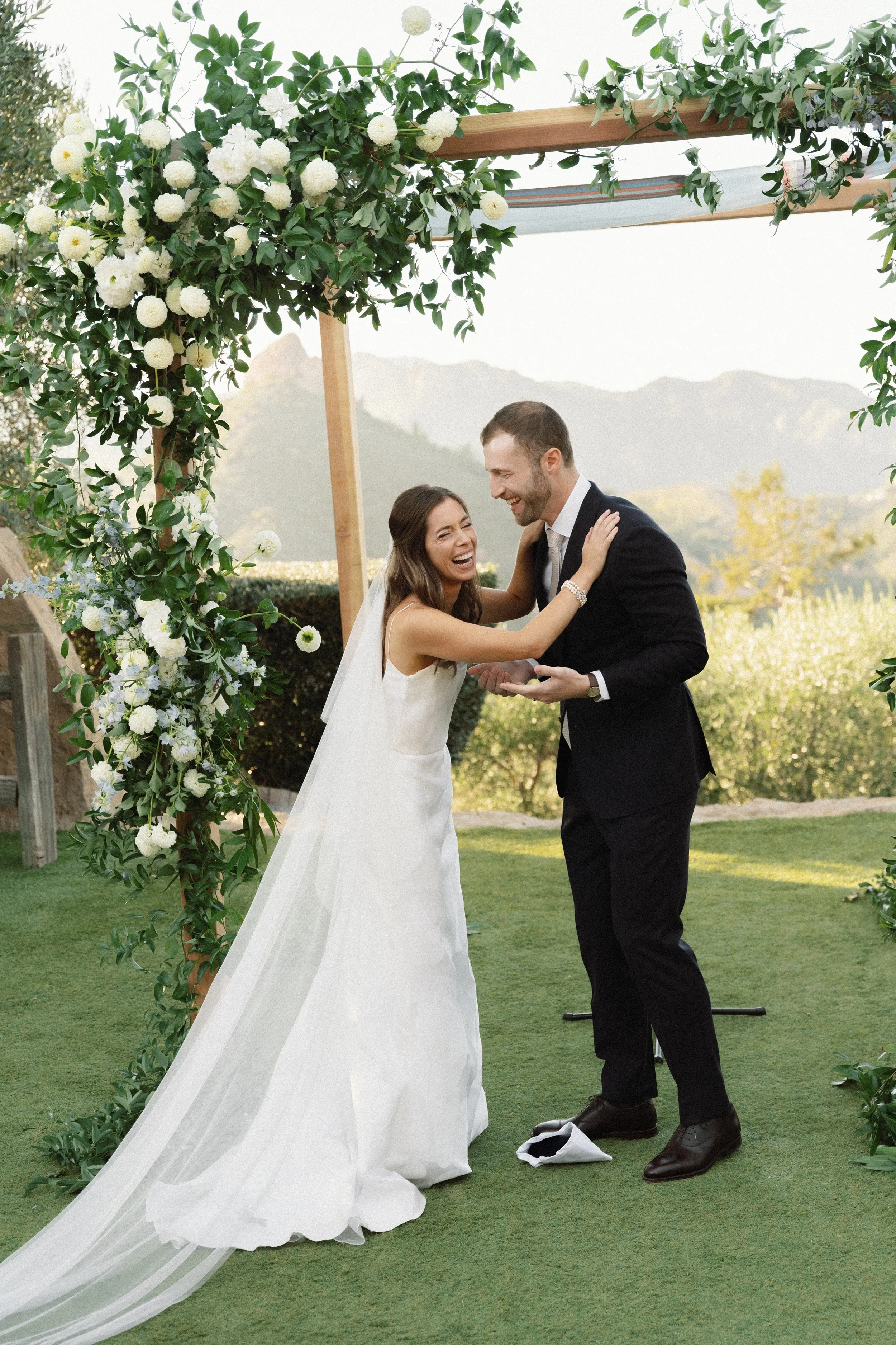 A bride and groom share a joyful moment during their outdoor wedding ceremony under a floral arch with green foliage and white flowers, mountains in the background.