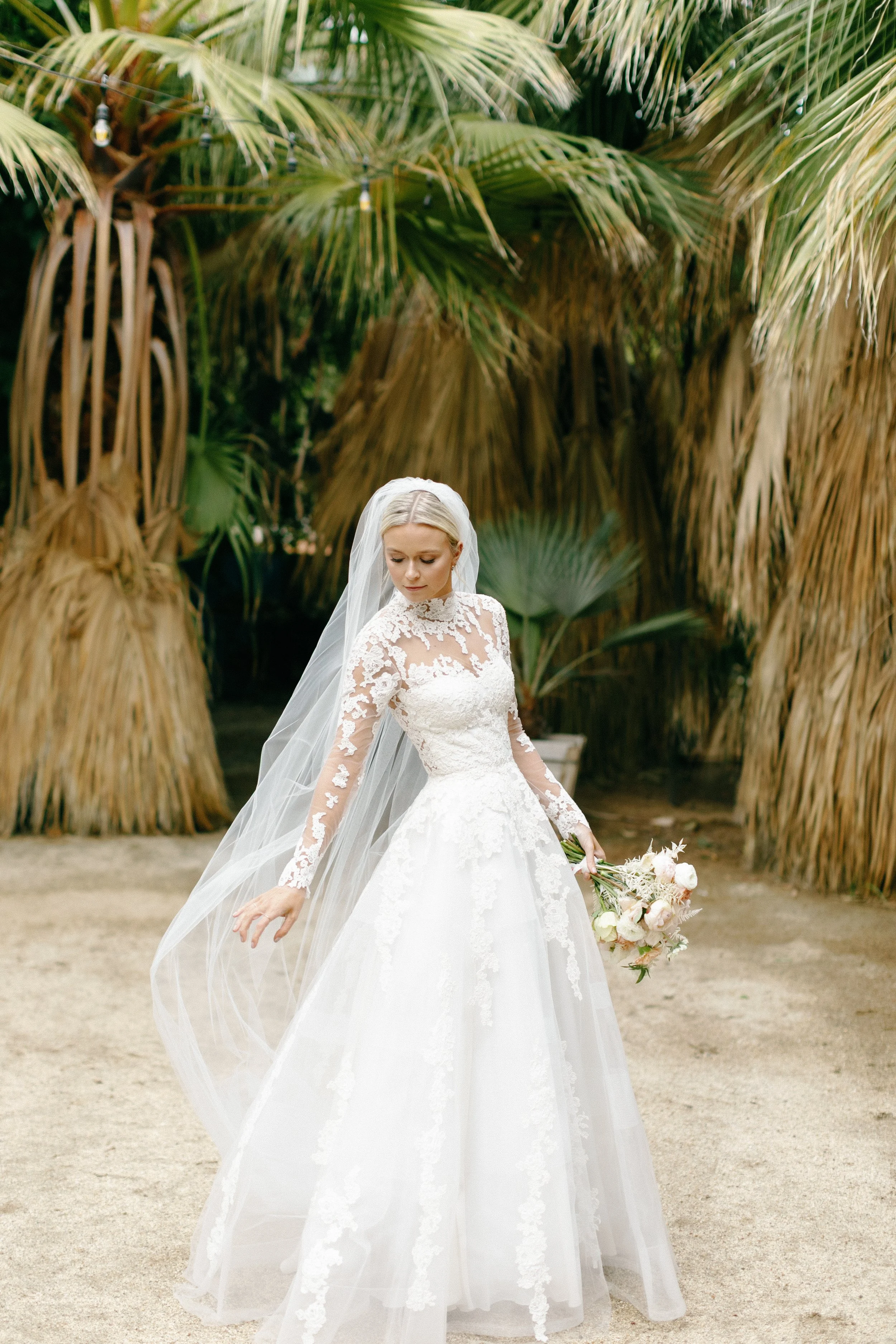 A bride in a white lace wedding dress holding a bouquet of flowers, standing outdoors with palm trees in the background.