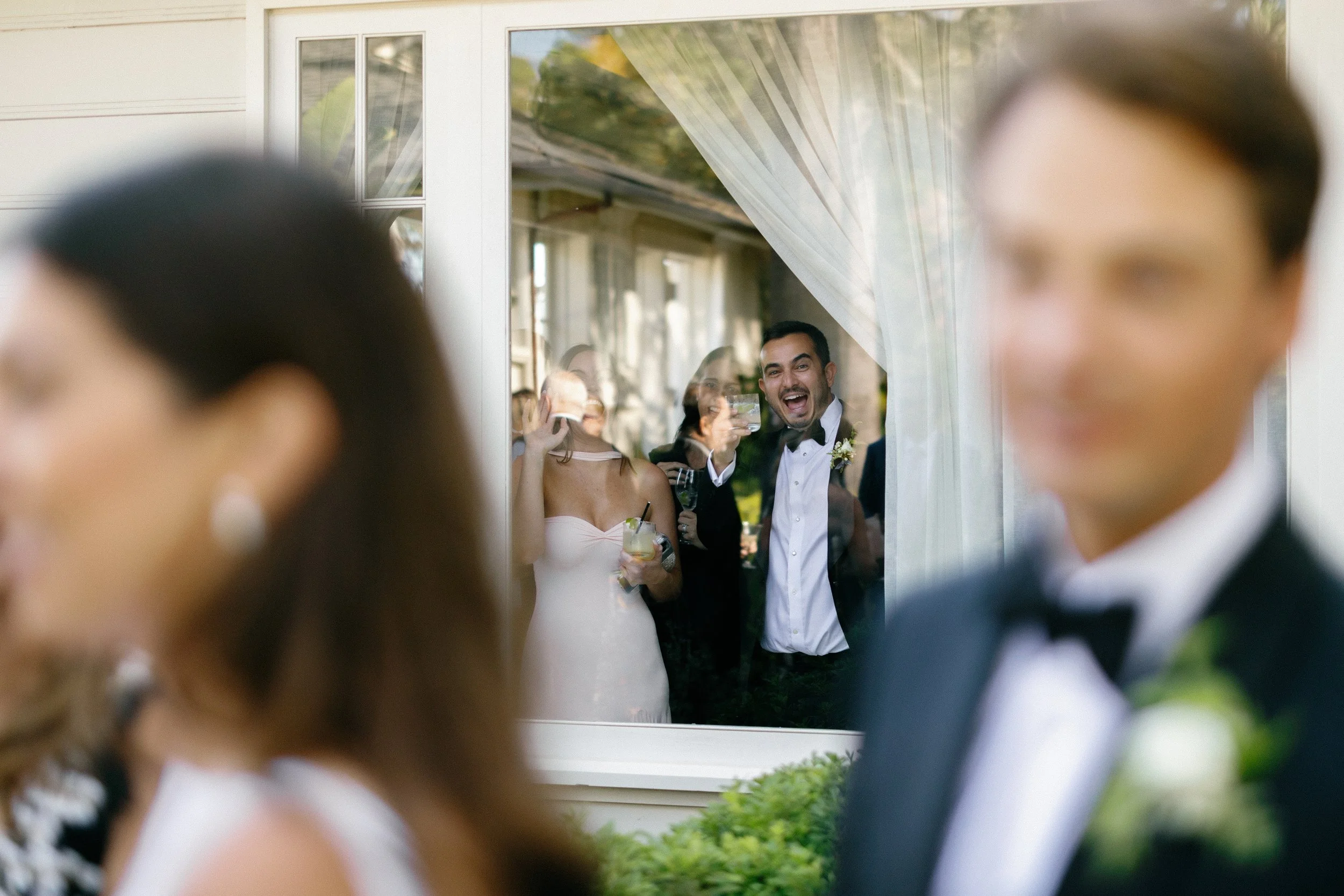 A group of people at a wedding, viewed through a window. Inside the window, a man in a tuxedo with a boutonniere cheers with a glass, smiling. Surrounding him are other guests holding drinks, with some wearing formal attire. The foreground shows two blurred people, likely part of the wedding party, with the focus on the joyful scene outside the window.