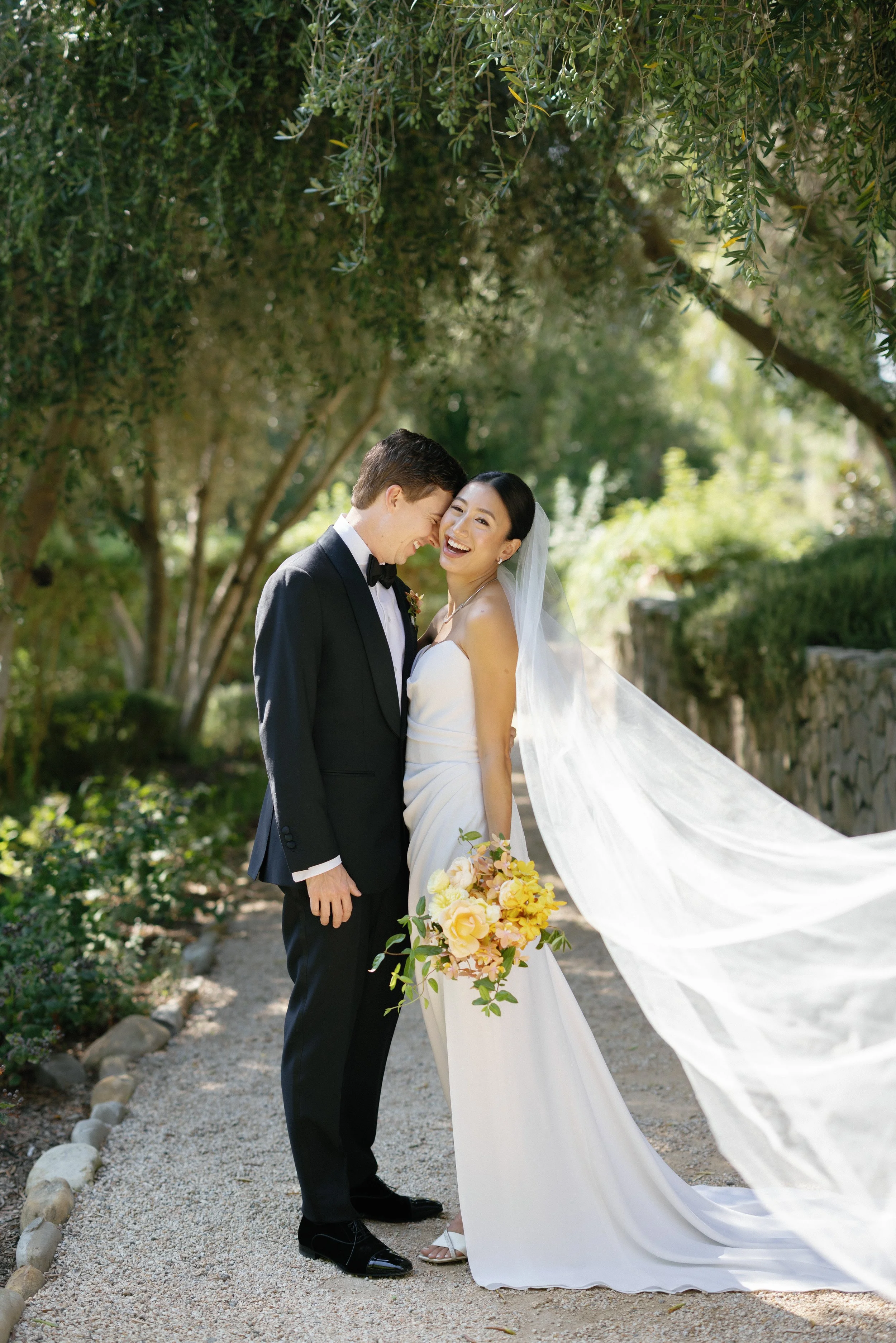 A joyful bride and groom sharing a moment outdoors during their wedding, with trees and greenery in the background, the bride holding a bouquet of yellow and pink flowers, and the bride in a white gown with a long veil.