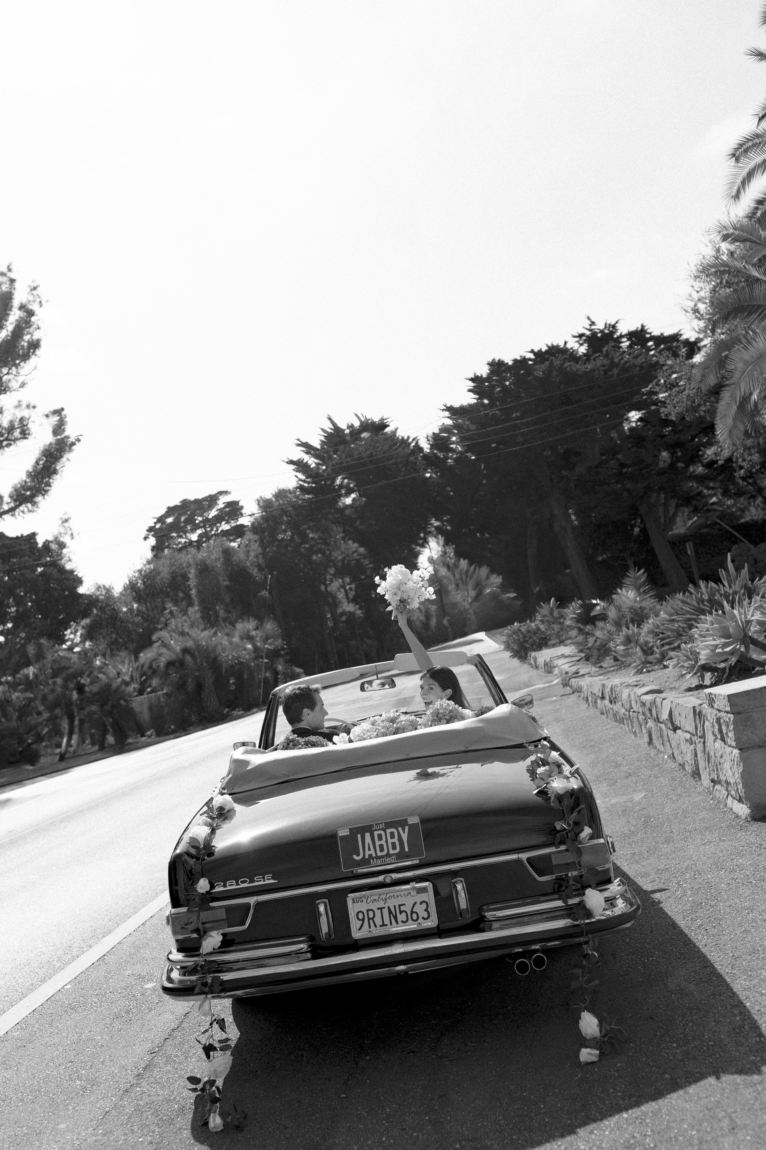 A black and white photo of a vintage car decorated with flowers, parked on the side of a road. Inside the car, a man and a woman are visible, with the woman holding a bouquet of flowers and the license plates read 9RIN563 and JABBY.