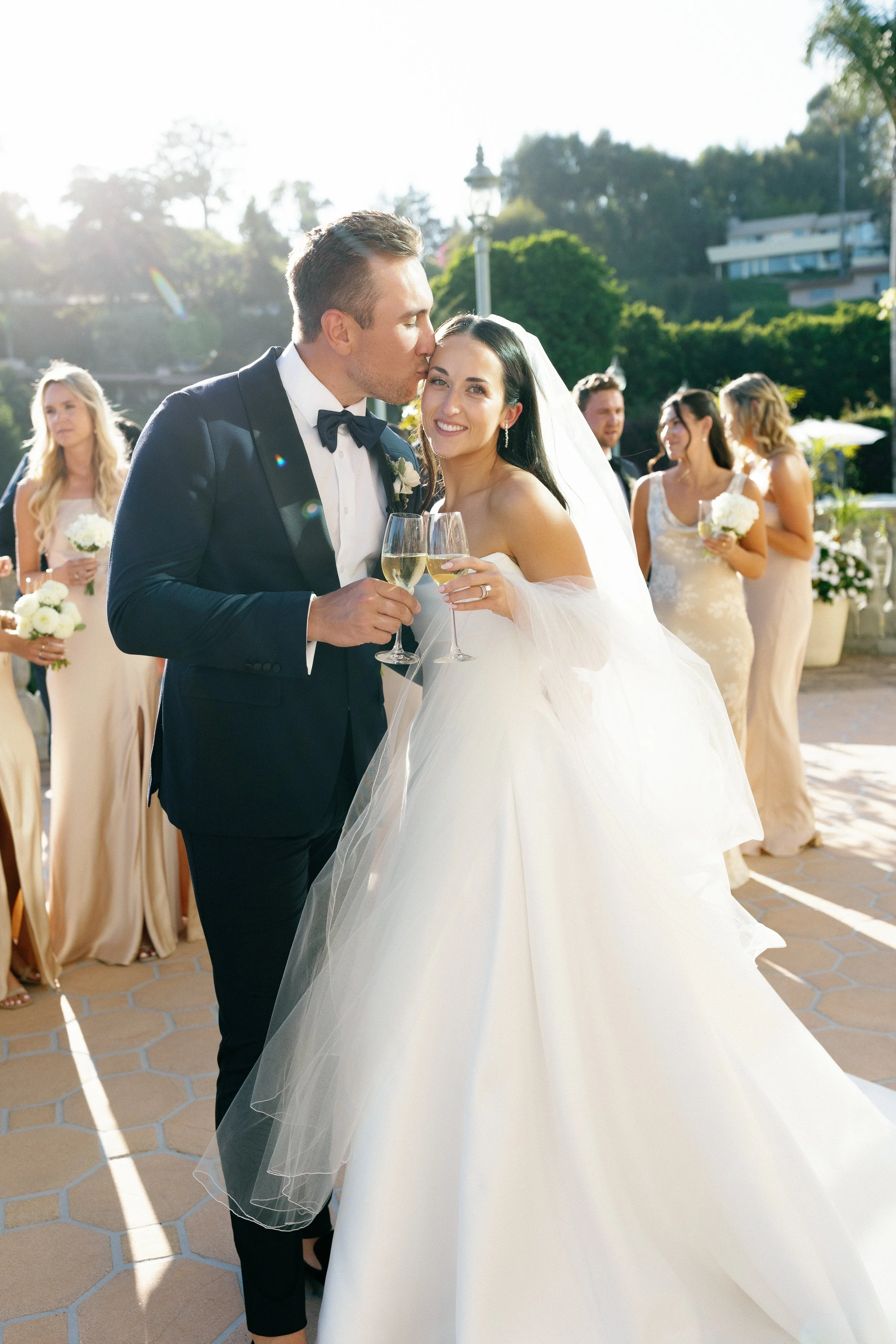 A newlywed couple at their wedding reception, standing outdoors with the groom kissing the bride on the forehead and both holding champagne glasses, surrounded by bridesmaids in beige dresses and greenery.