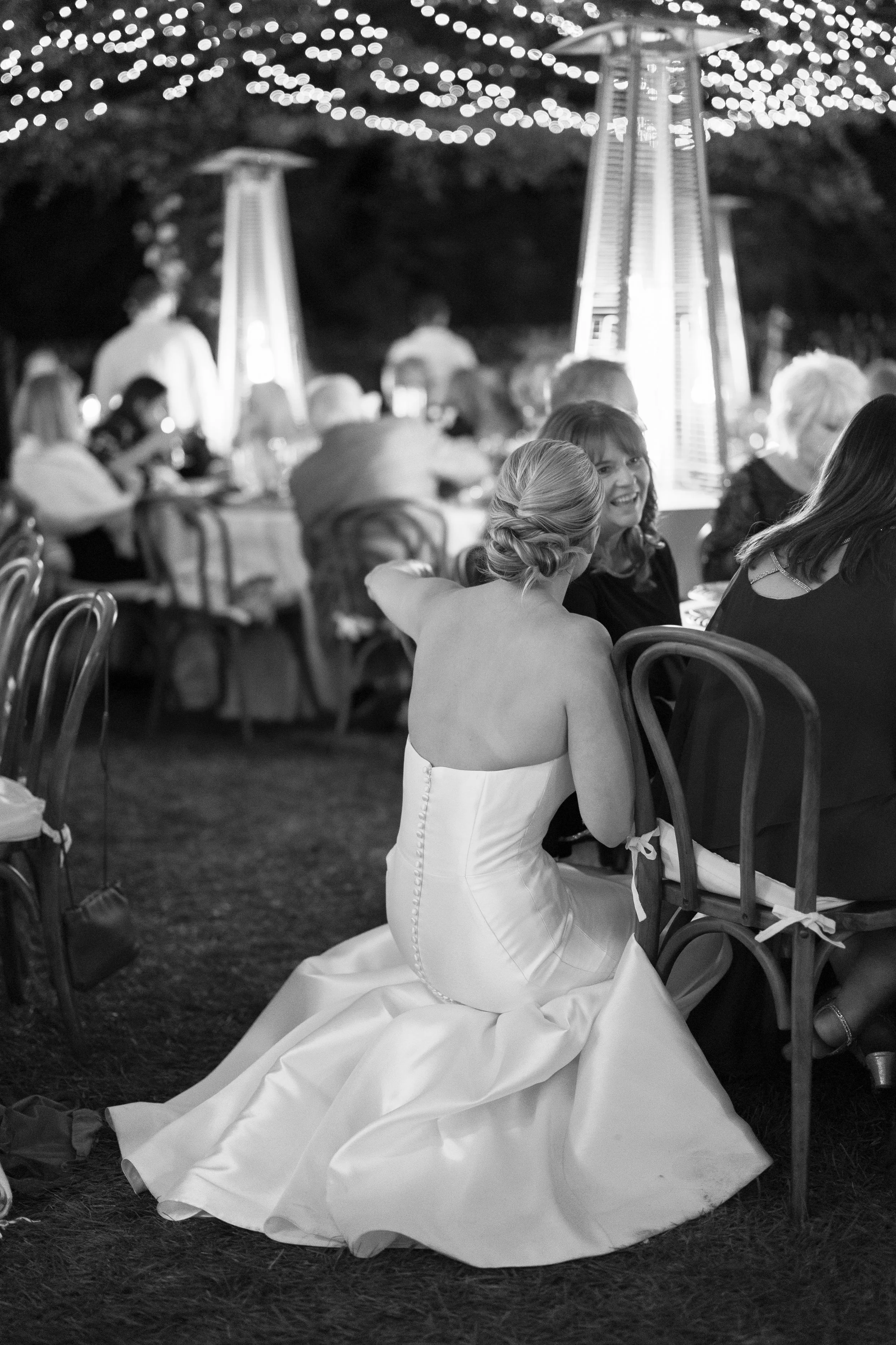 A woman in a strapless wedding gown sitting on the ground at an outdoor evening event, surrounded by seated guests and string lights.