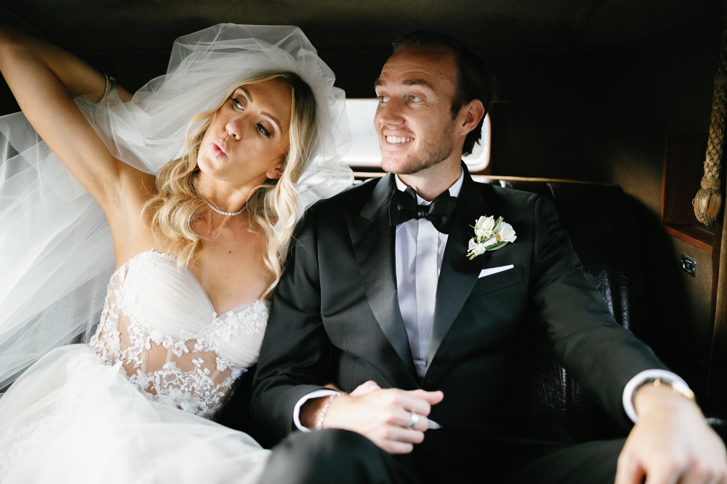 A bride and groom sitting inside a vehicle, dressed in wedding attire, with the bride making a playful face and the groom smiling.