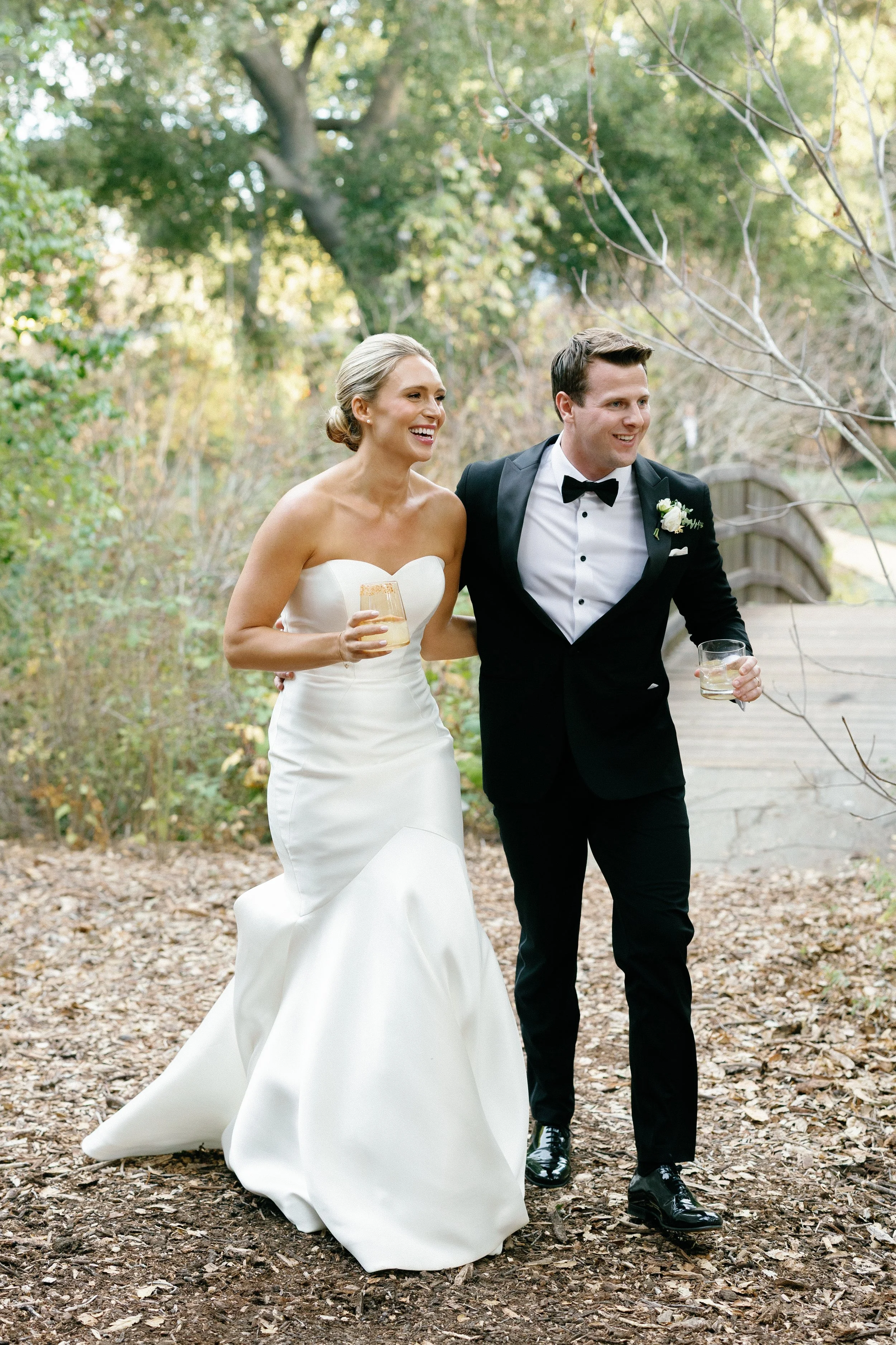A bride and groom walking outdoors on a wooded path, smiling, holding drinks, and dressed in wedding attire.