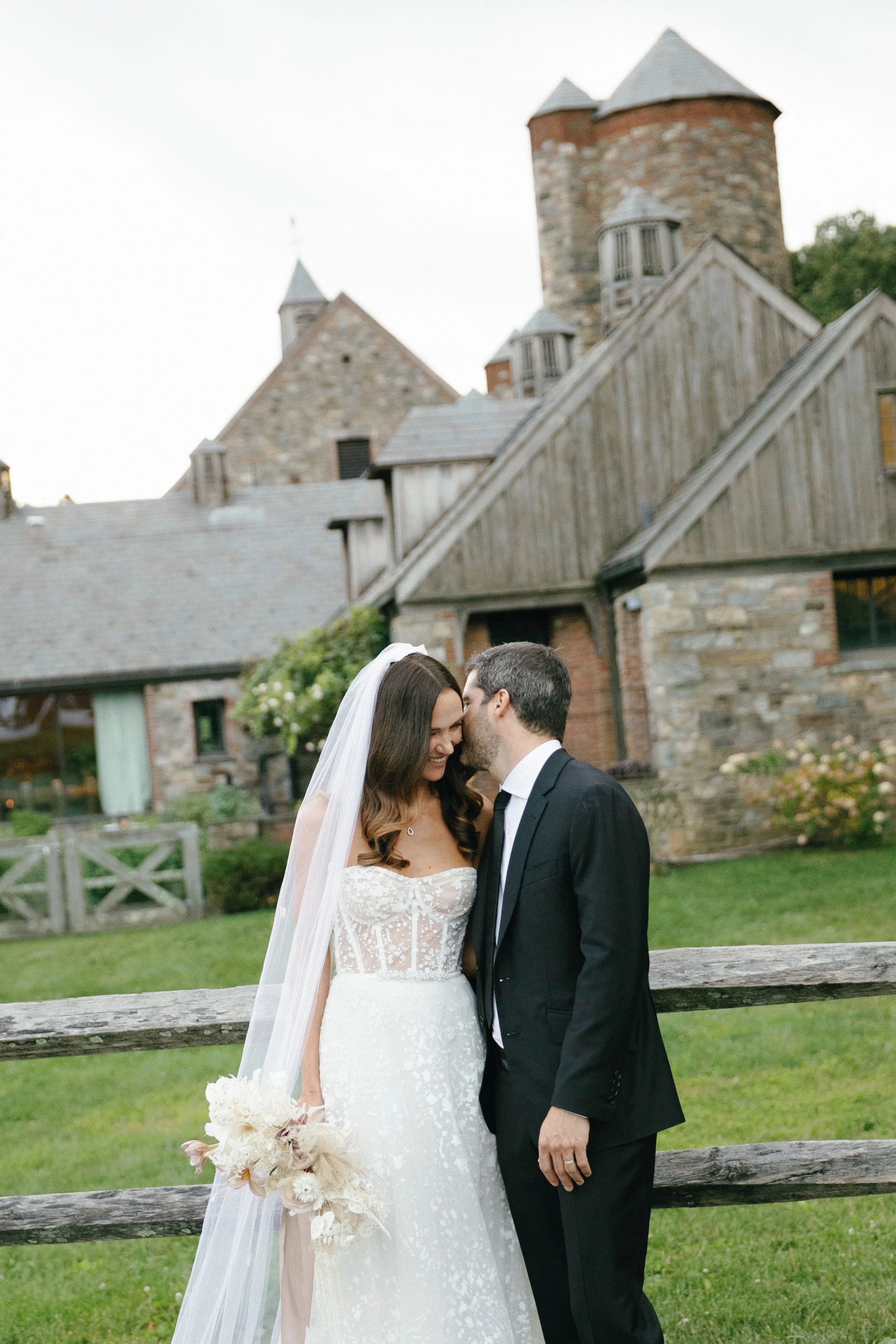 A bride and groom share a kiss outdoors in front of a rustic stone and wood building, with grass and a wooden fence in the background.