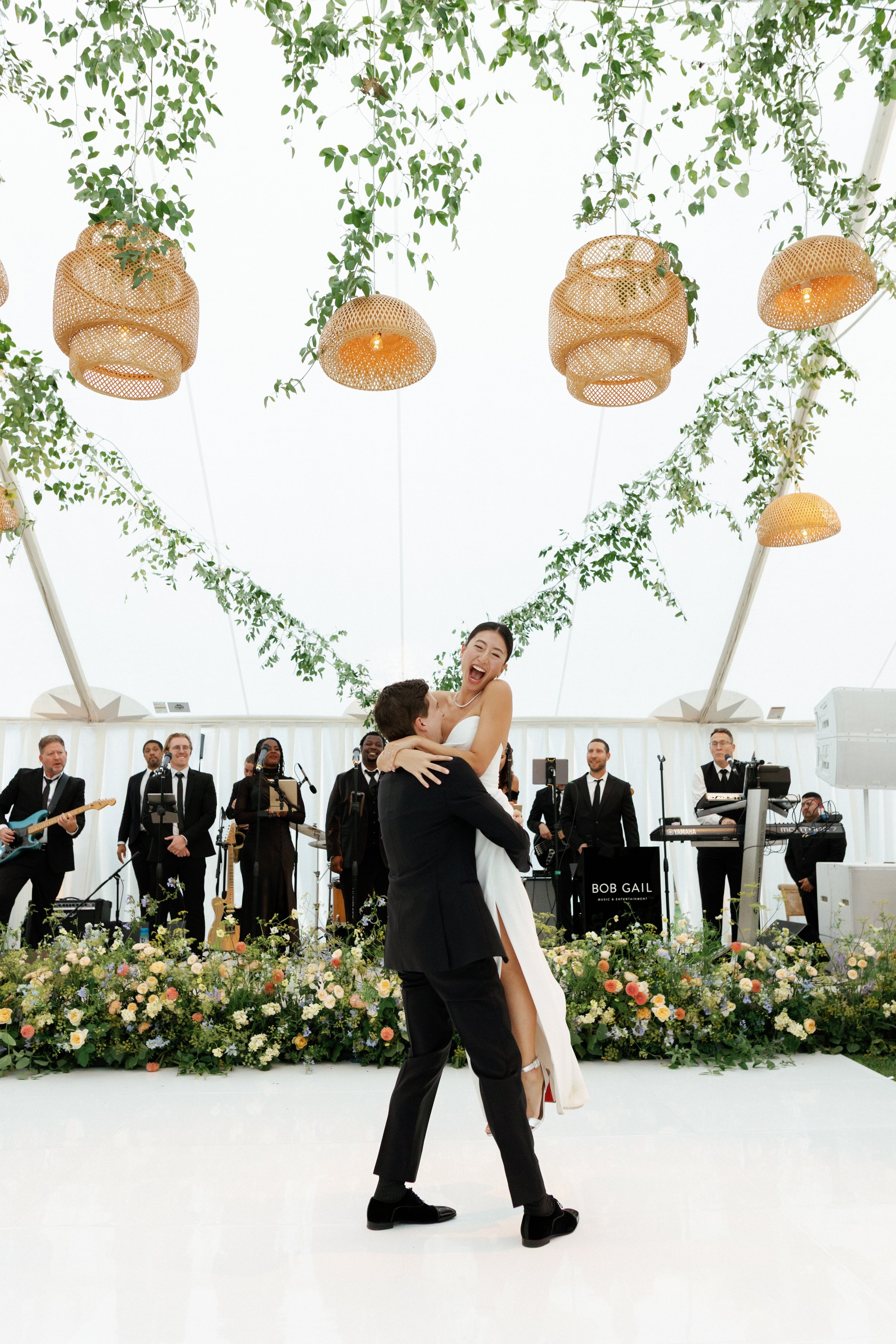 A groom lifting and kissing a bride during a wedding reception or celebration, with a band playing in the background and floral decorations.