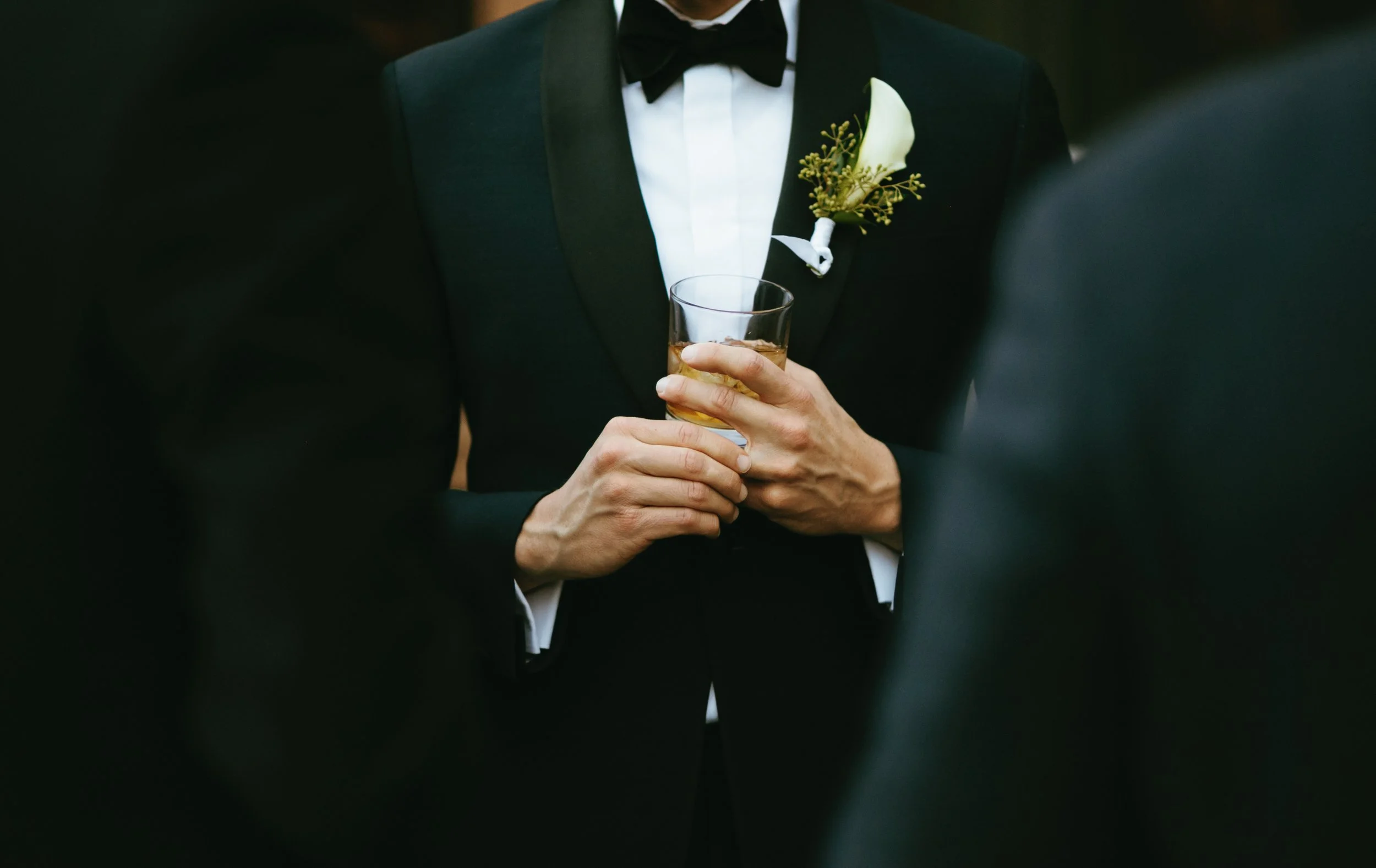 Man in a black tuxedo holding a glass of whiskey at a formal event.