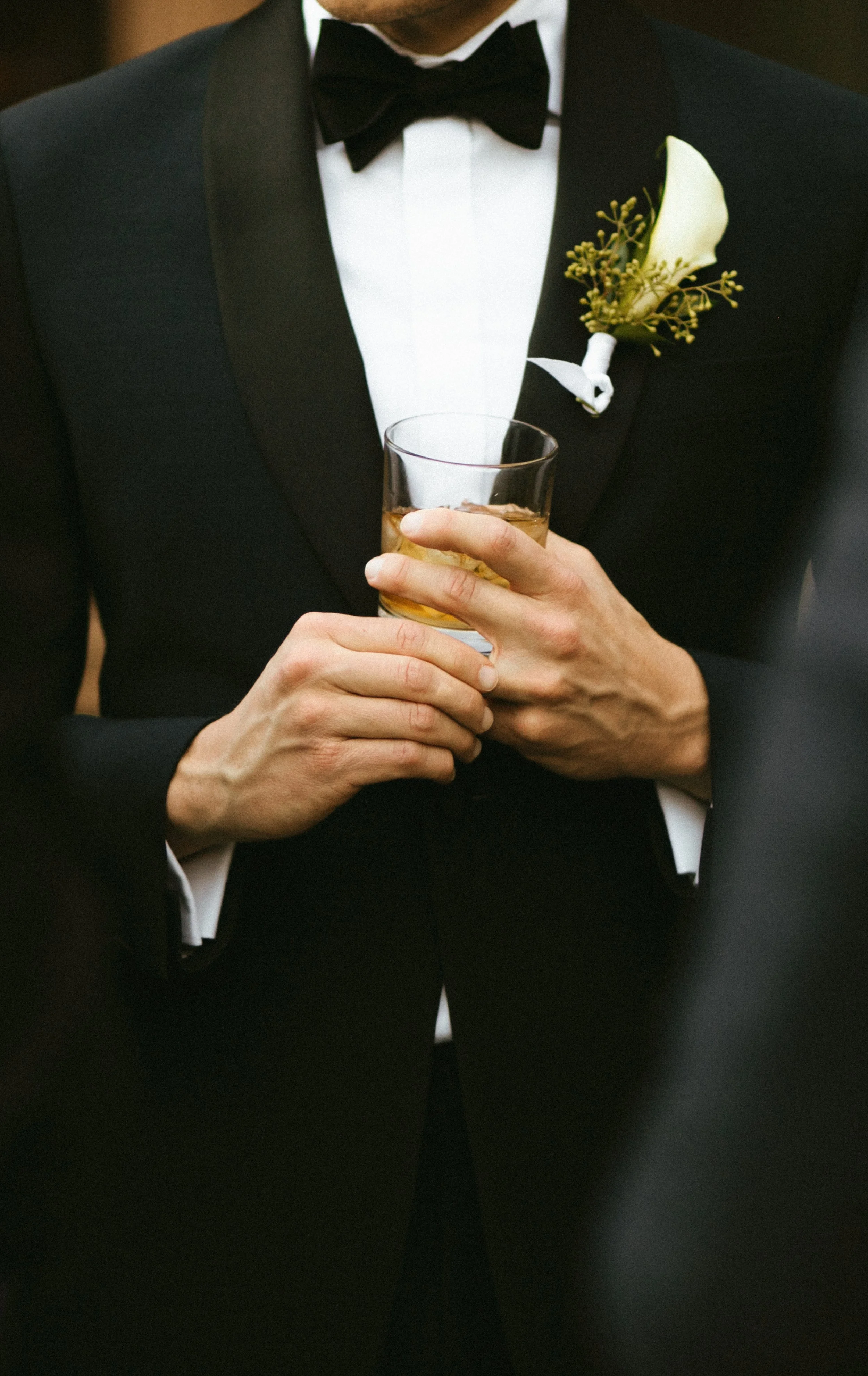 A man in a tuxedo holding a glass of whiskey at a formal event.