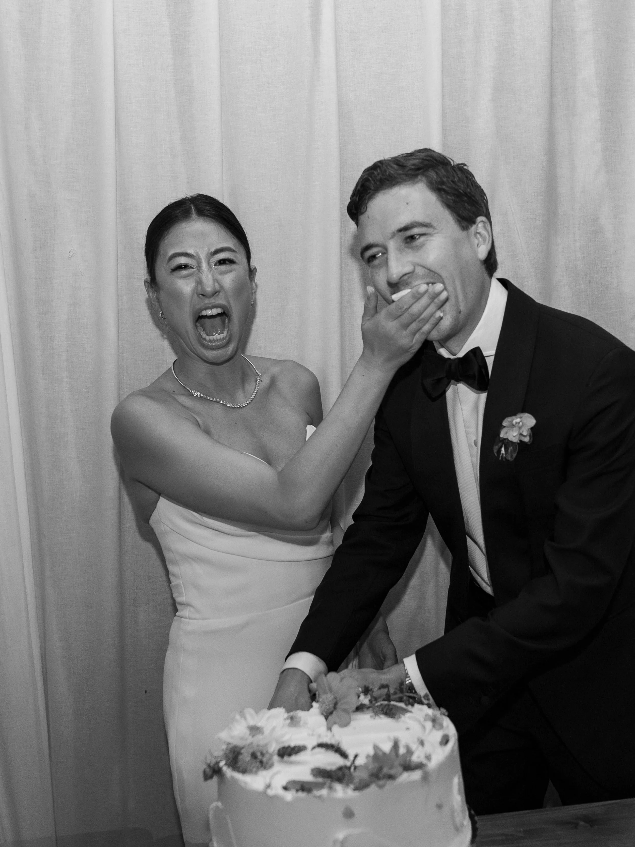 A woman in a wedding dress playfully smears cake on a groom's face during a wedding reception, with a cake decorated with flowers in front of them.