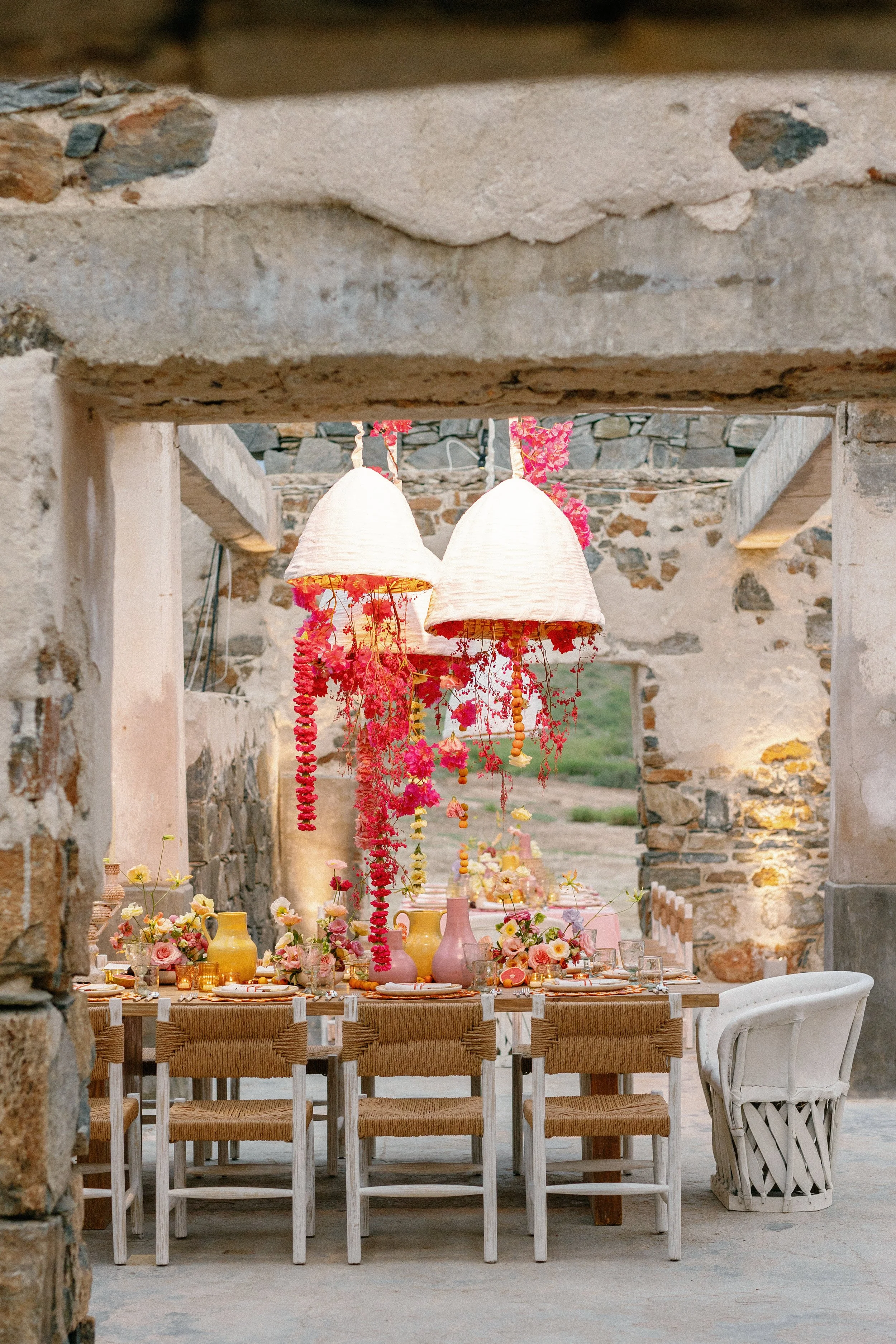 A rustic outdoor dining setup with a wooden table and chairs decorated with pink and white flowers. Hanging from the ceiling are two large white lampshades adorned with pink and red floral garlands. The setting is in a stone-walled courtyard with a s