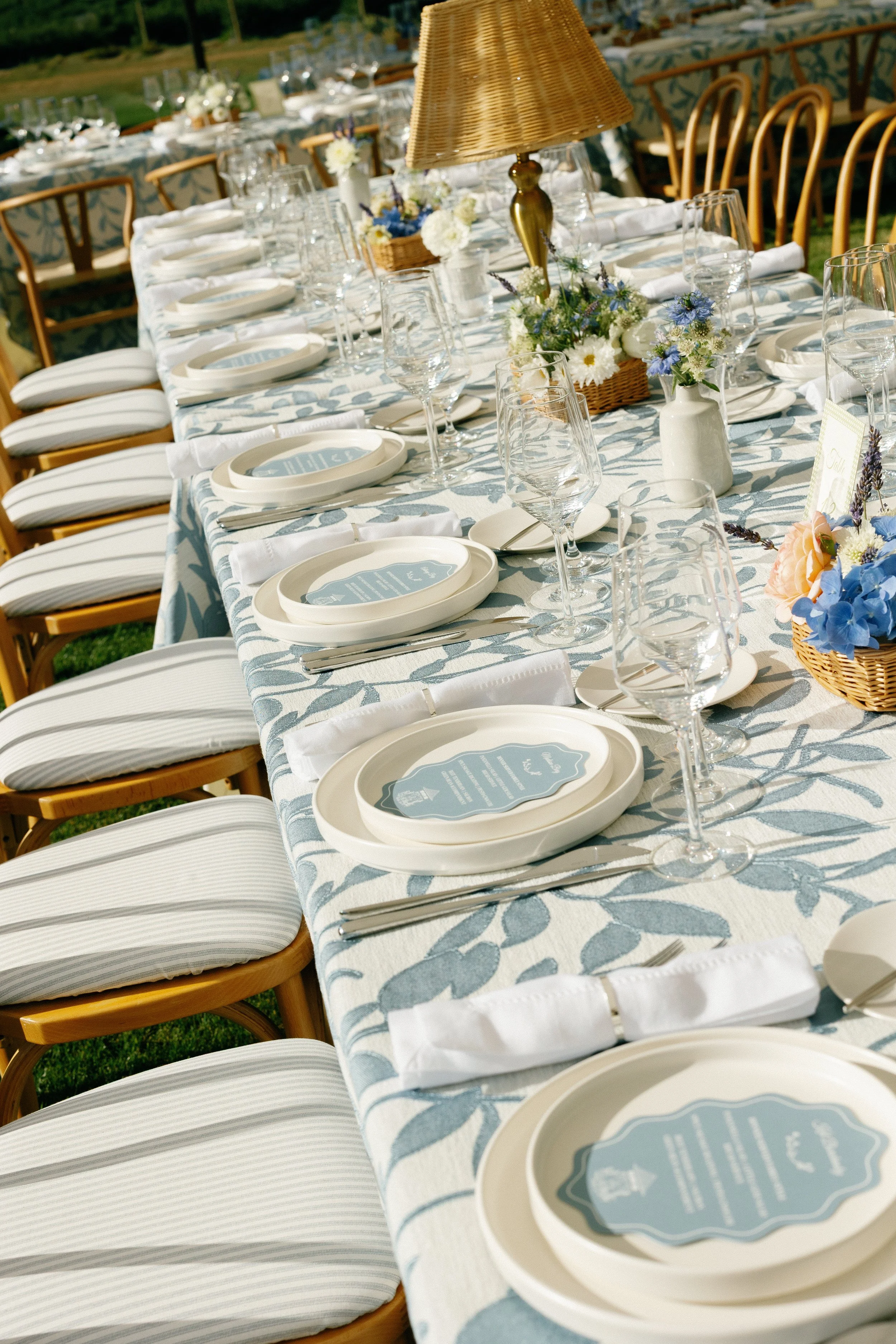 Outdoor banquet table decorated with white and blue tablecloth, floral centerpieces, white plates, napkins, silverware, and glassware, set for a formal event.