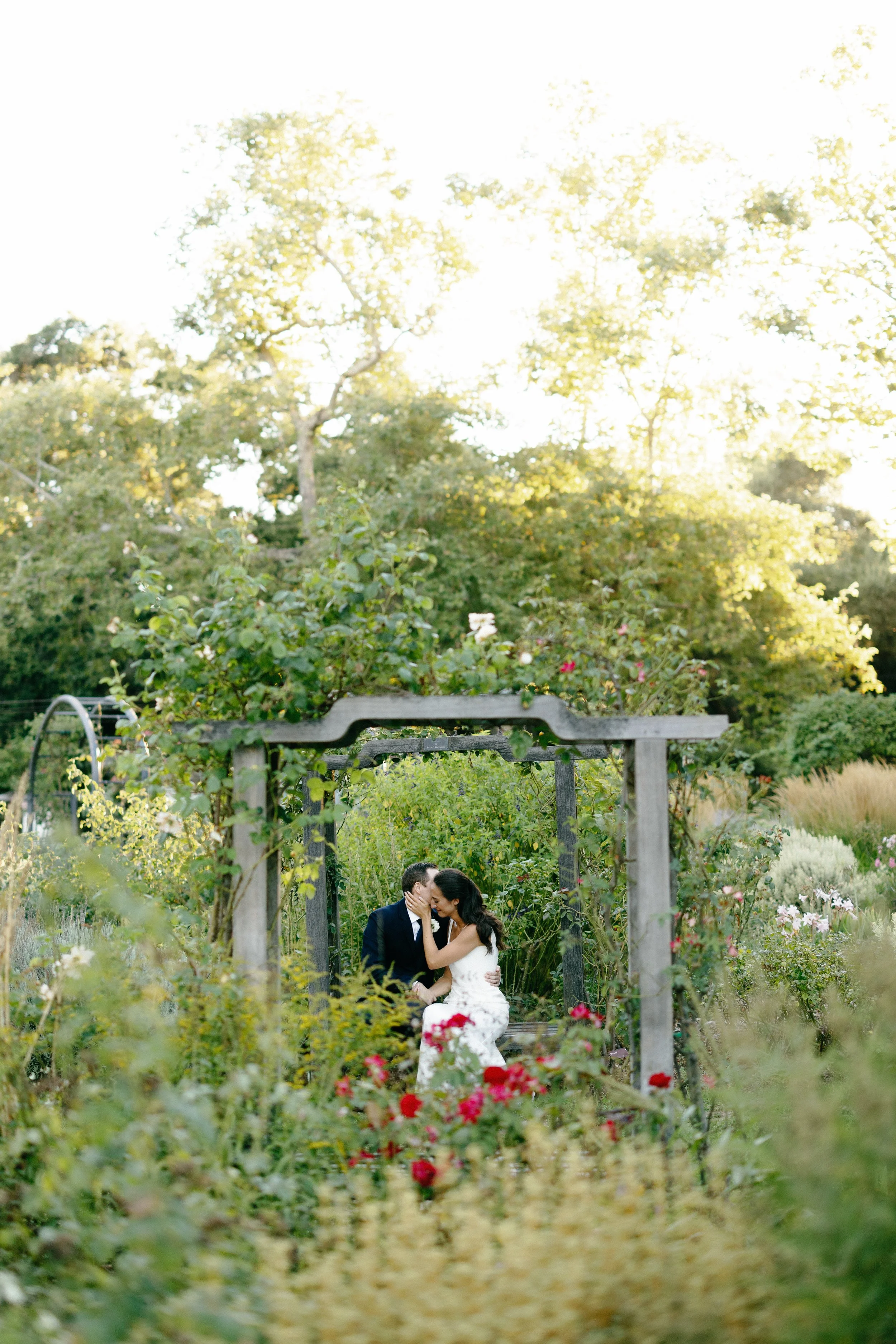 A couple dressed in wedding attire sitting close together in a lush garden with blooming flowers, sharing an intimate moment under a wooden archway during the daytime.