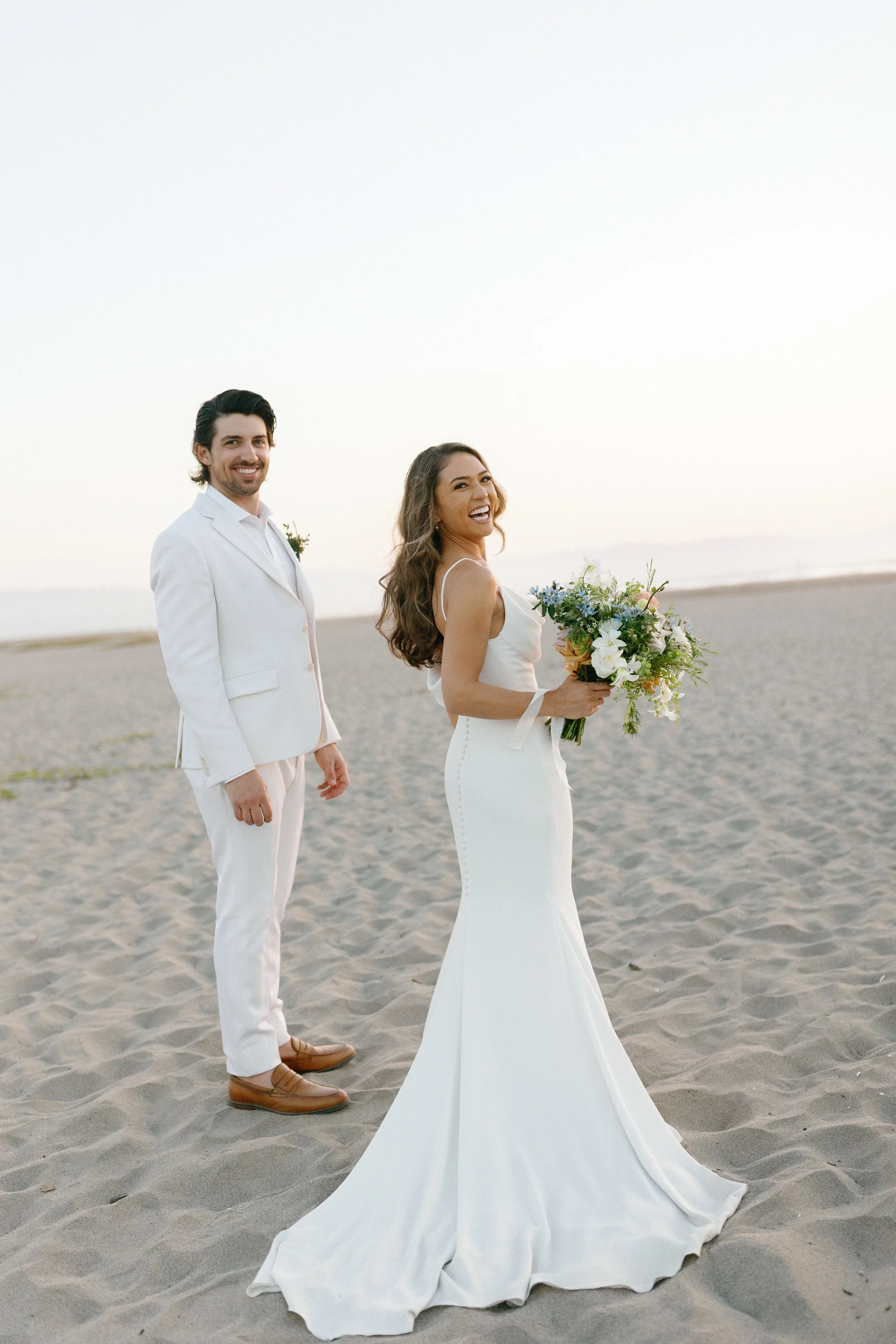 A bride in a white dress holding a bouquet of flowers and a groom in a white suit standing on a sandy beach at sunset, both smiling.