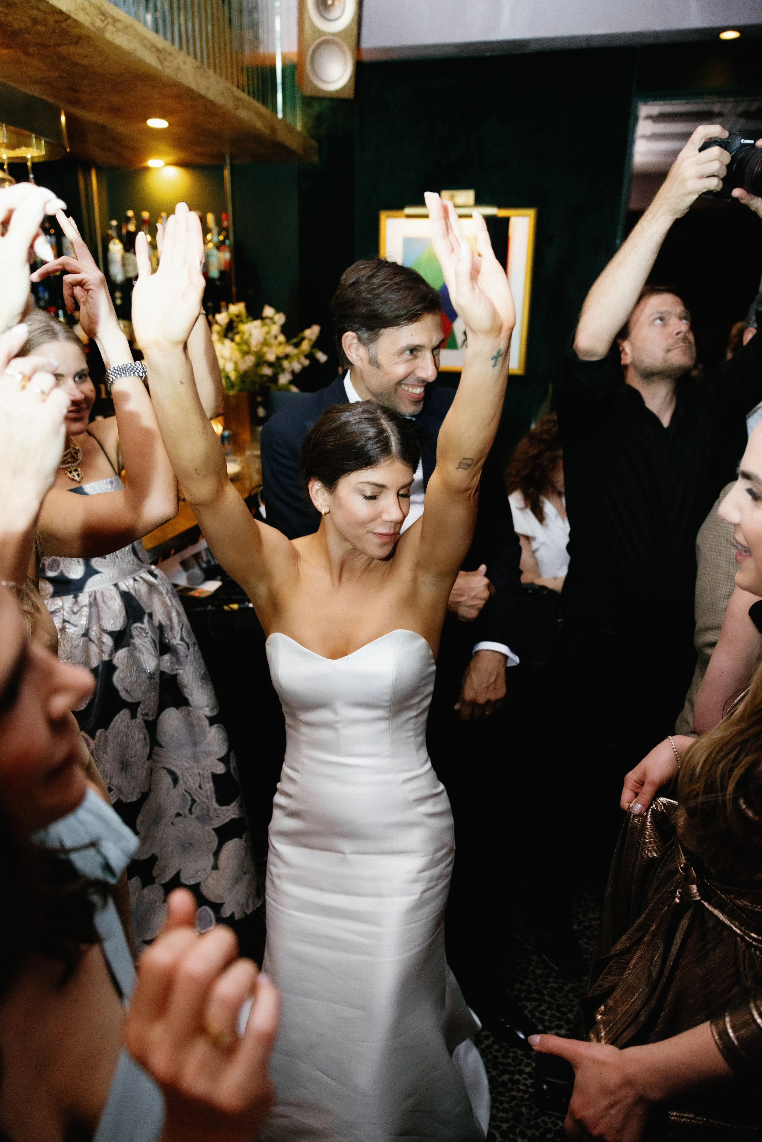 A bride in a strapless white wedding dress dancing with her arms raised at her wedding reception, surrounded by guests in celebration.