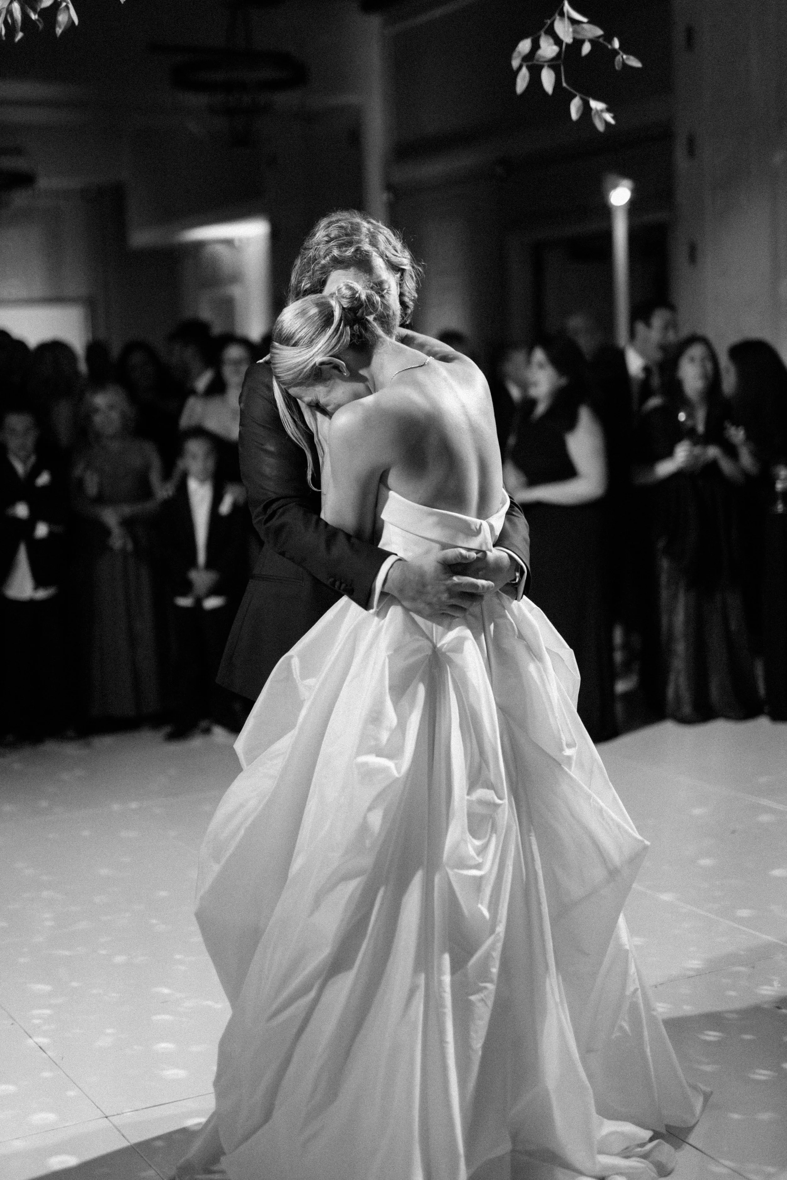 A bride and groom share a dance at their wedding reception, surrounded by guests in the background.