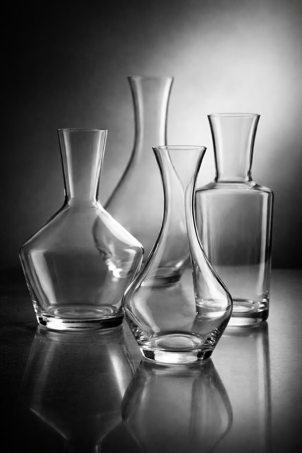 Four empty glass vases of varying shapes and sizes on a reflective surface with a dark gradient background.