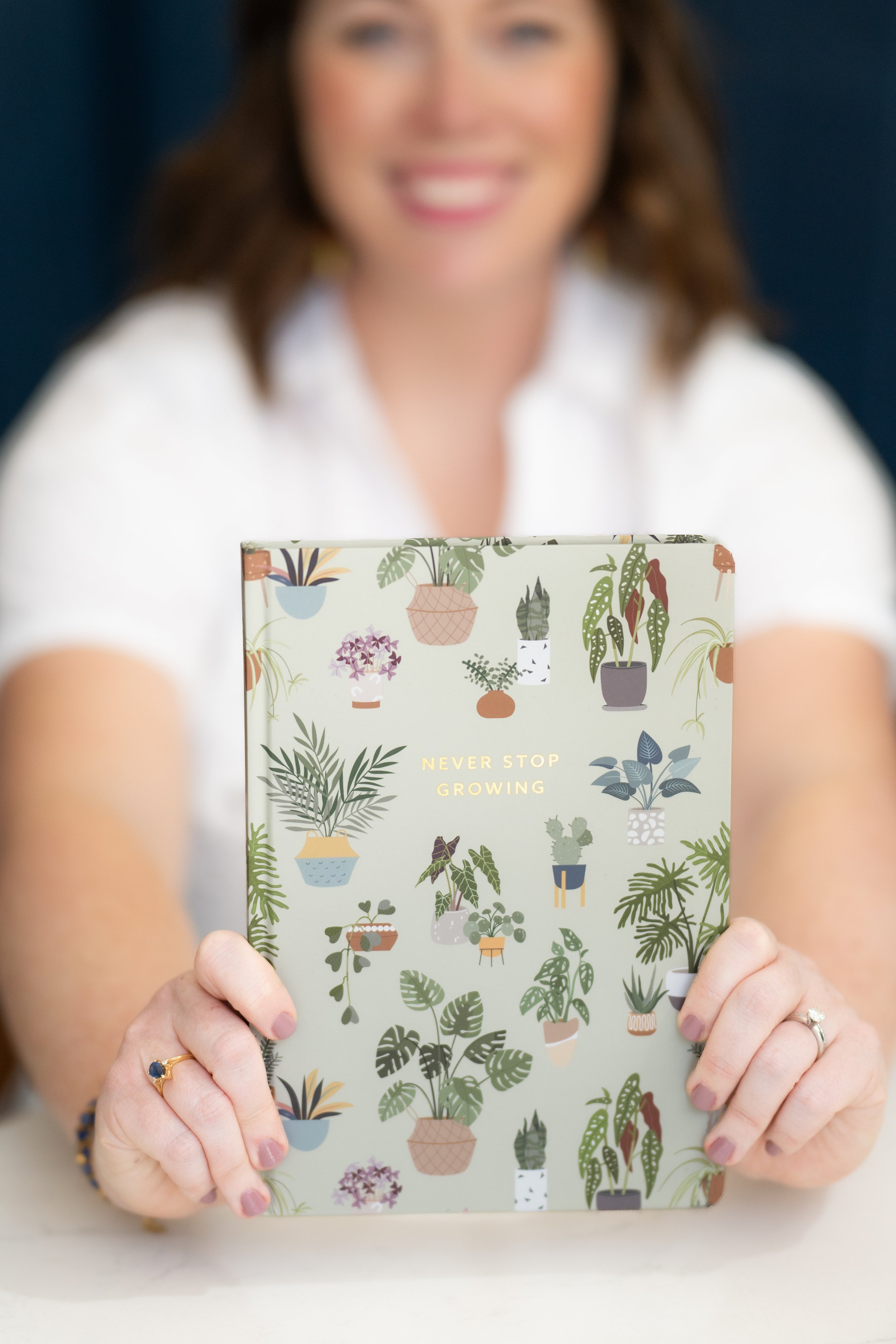 A woman holding a colorful illustrated book titled 'Never Stop Growing' featuring various potted plants and flowers on the cover.