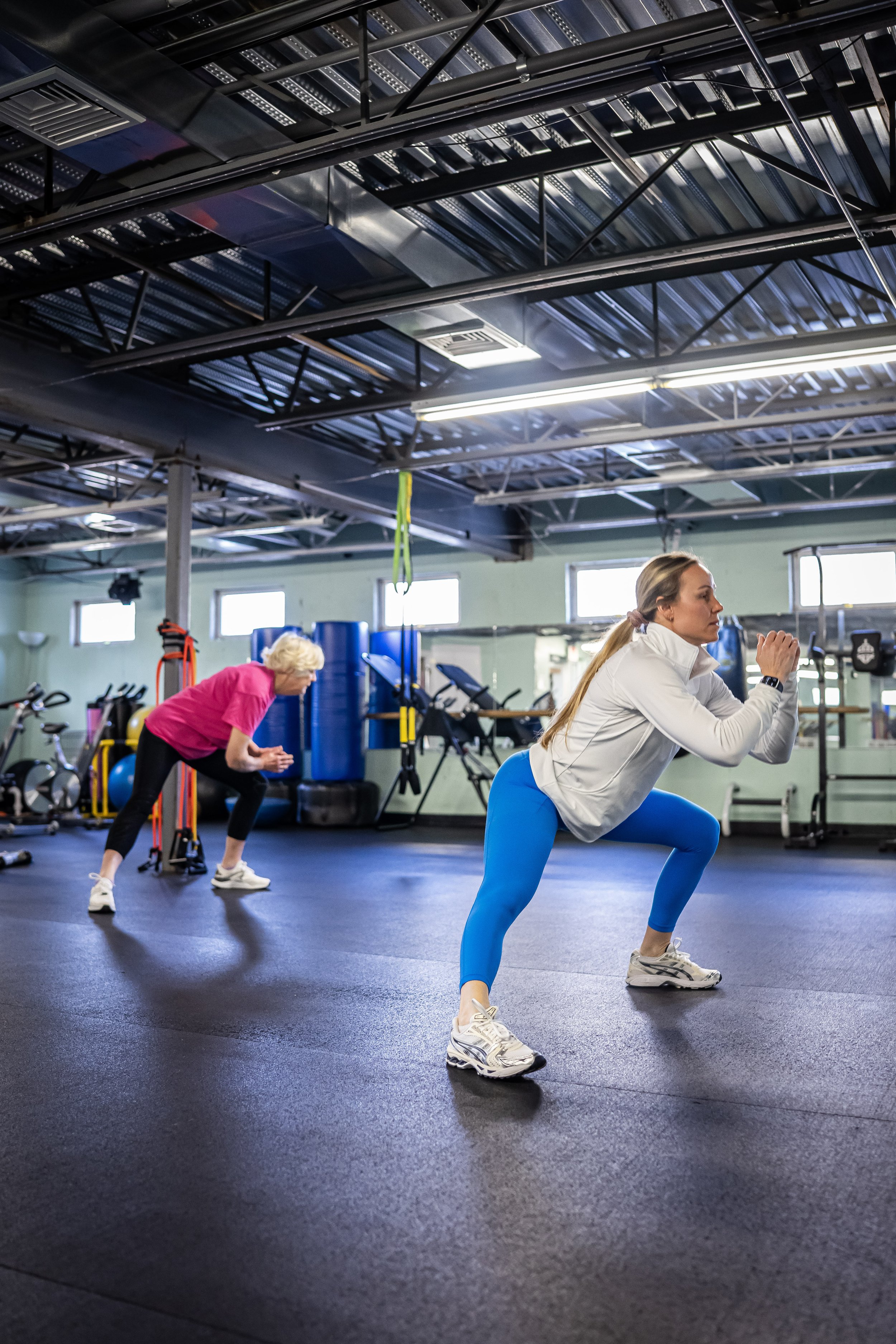 Two women exercising in a gym performing squats, one in blue leggings and a white top, the other in a pink top and black pants.