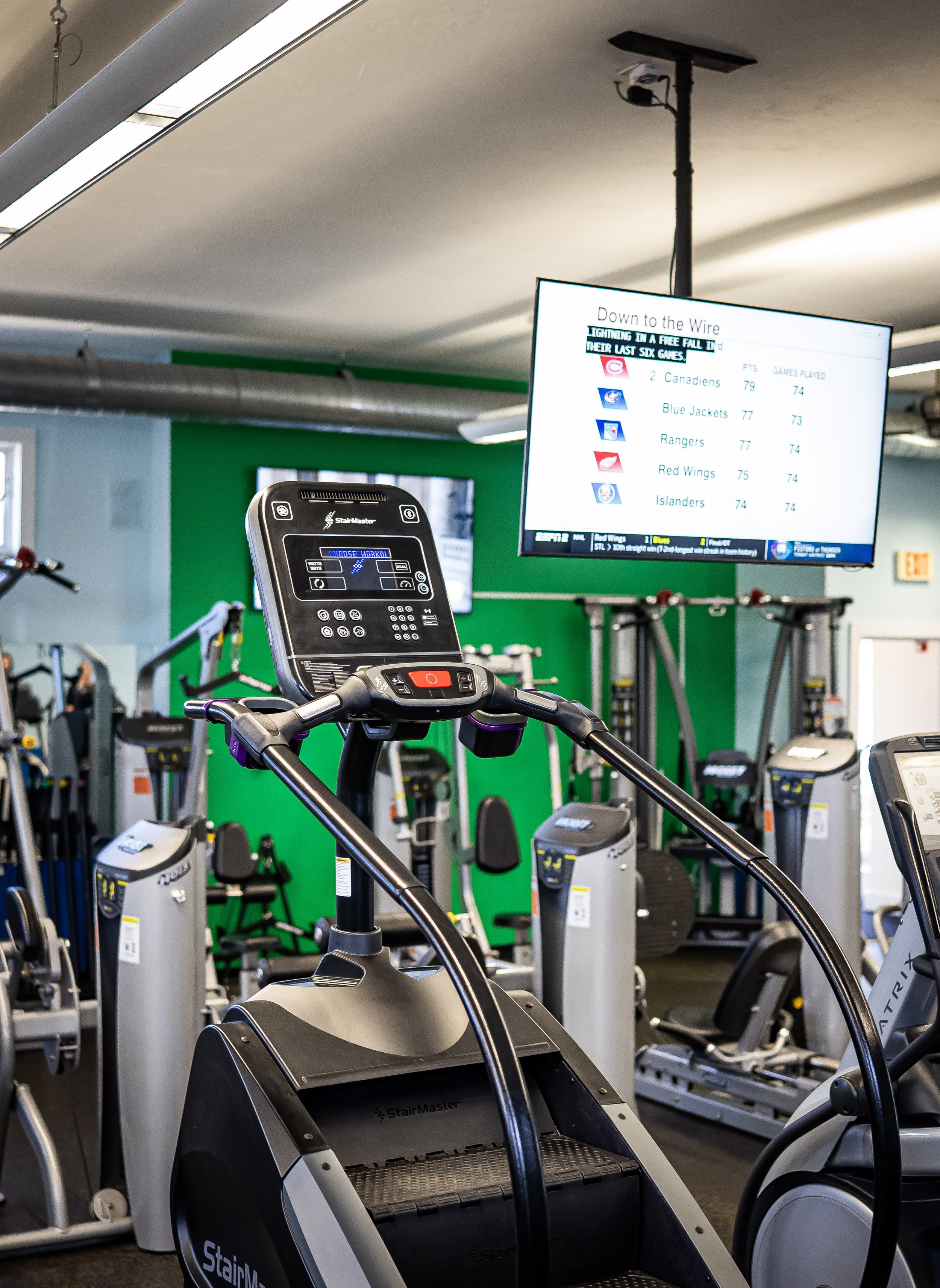 Gym interior with exercise machines, including a StairMaster in the foreground and a television displaying sports standings on the wall.