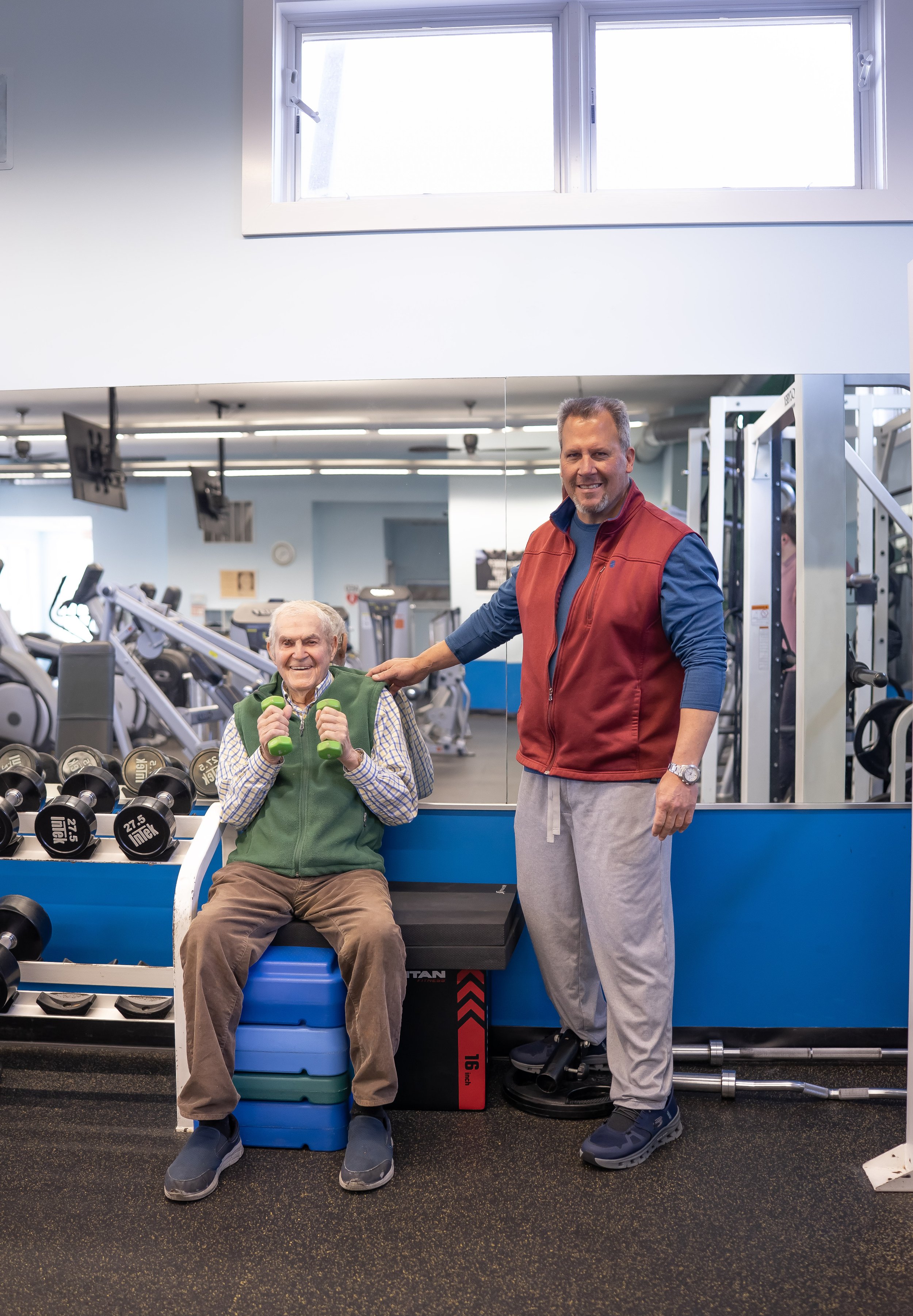 An elderly man holding green dumbbells sits on a stack of exercise steps in a gym, smiling. A middle-aged man in a red vest, gray sweatpants, and watch stands beside him, also smiling. The gym has various exercise equipment, including dumbbells and a