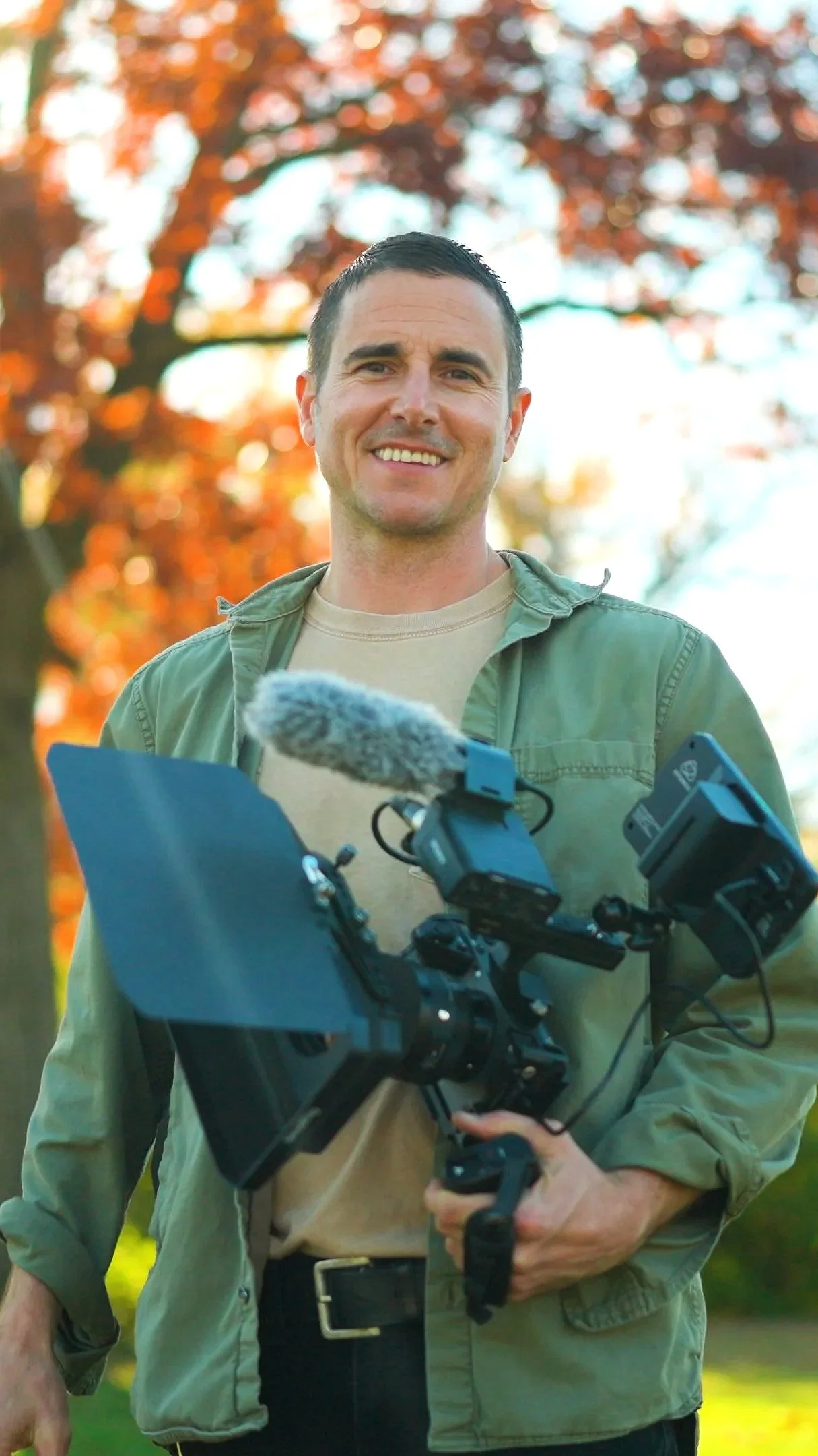 Man holding professional video camera outdoors in front of a tree with orange and red autumn leaves, smiling at the camera.