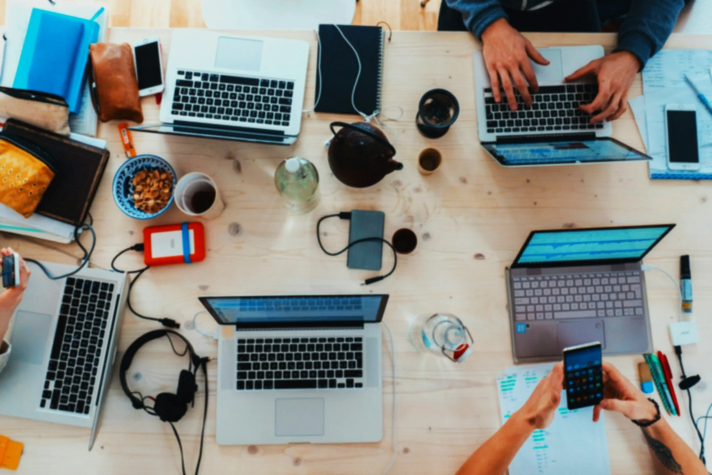 Overhead view of a cluttered wooden table with six people working on laptops, surrounded by notebooks, phones, a water bottle, a teapot, cups, pens, and snacks.