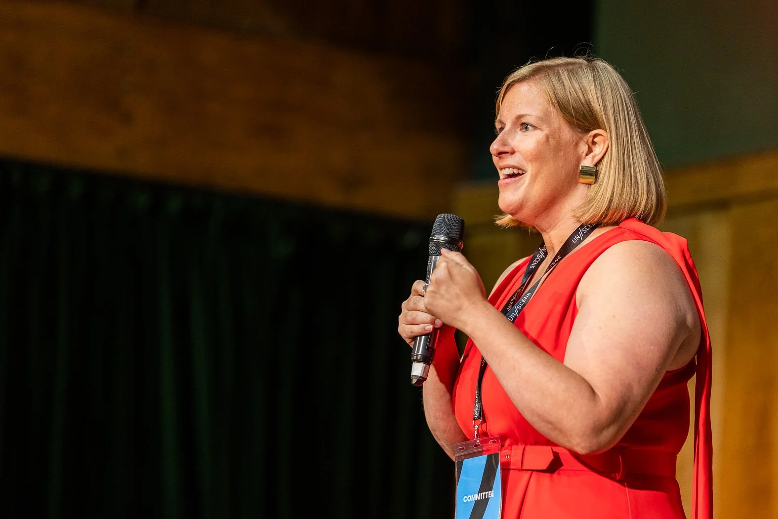 Lucy Barbor, our founder, on stage giving the welcome speech for the first UN//SCENE festial. Lucy wears a red jumpsuit and is holding a microphone, looking justifiably proud and delighted.