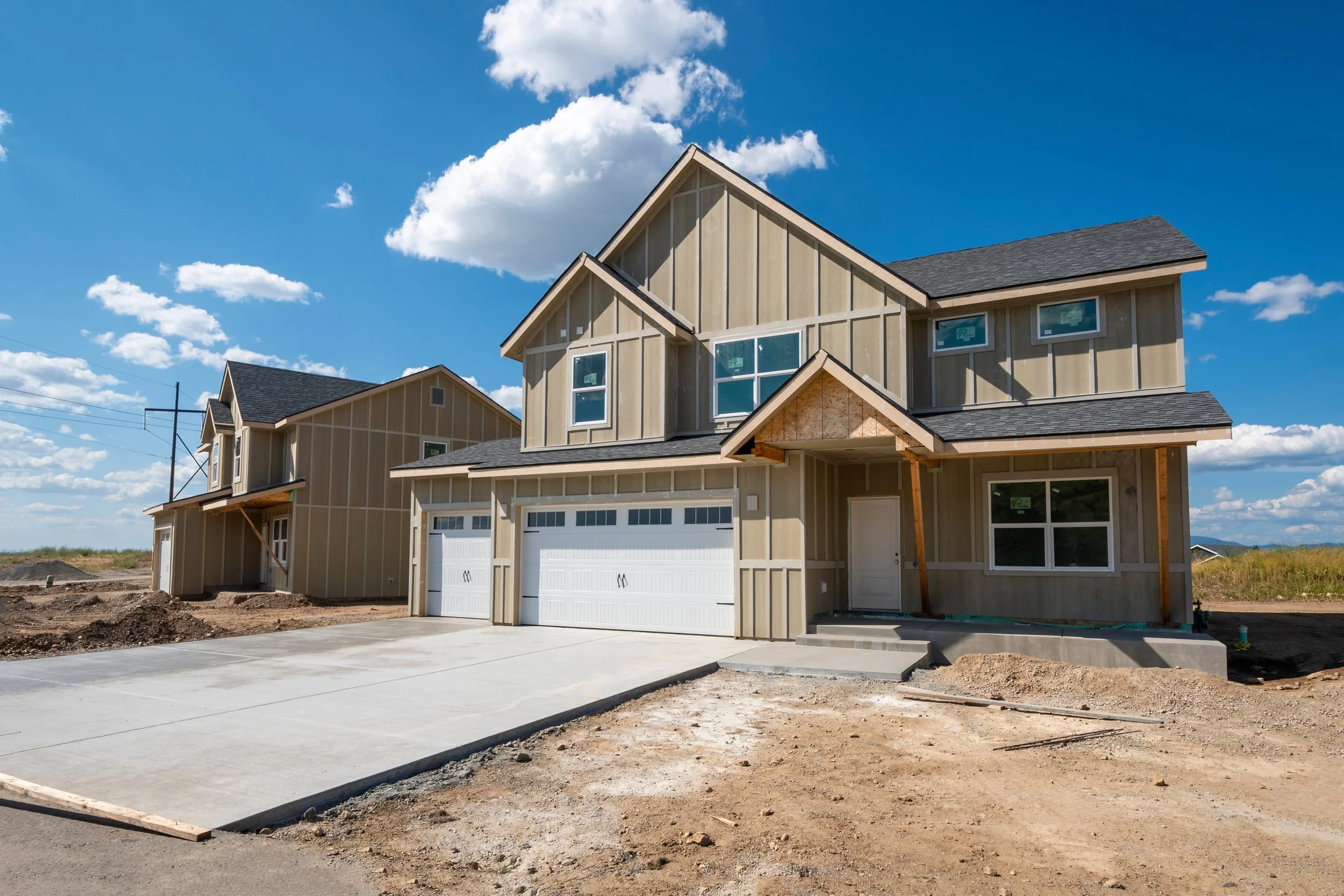 Newly built two-story house under construction with a concrete driveway, multiple windows, and a gray shingled roof, against a bright blue sky with scattered clouds.