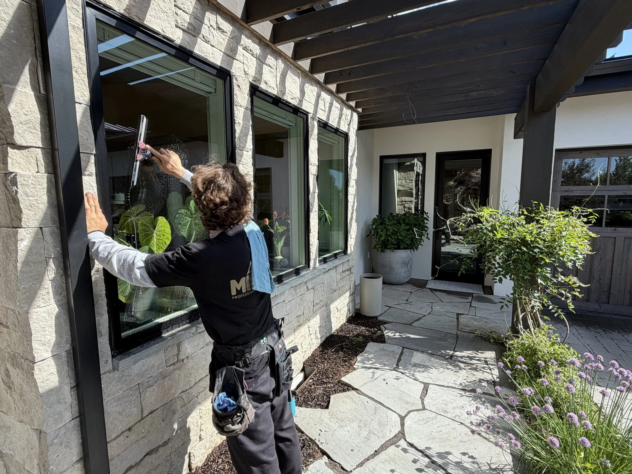 A person cleaning a large window on a stone exterior wall of a modern house, with plants and flowers in the garden nearby in Bend, Oregon.