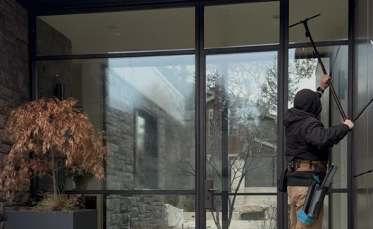 A person wearing black hooded jacket and carrying tools is cleaning a large glass window using a squeegee inside a modern building with a brick wall and a potted plant nearby.