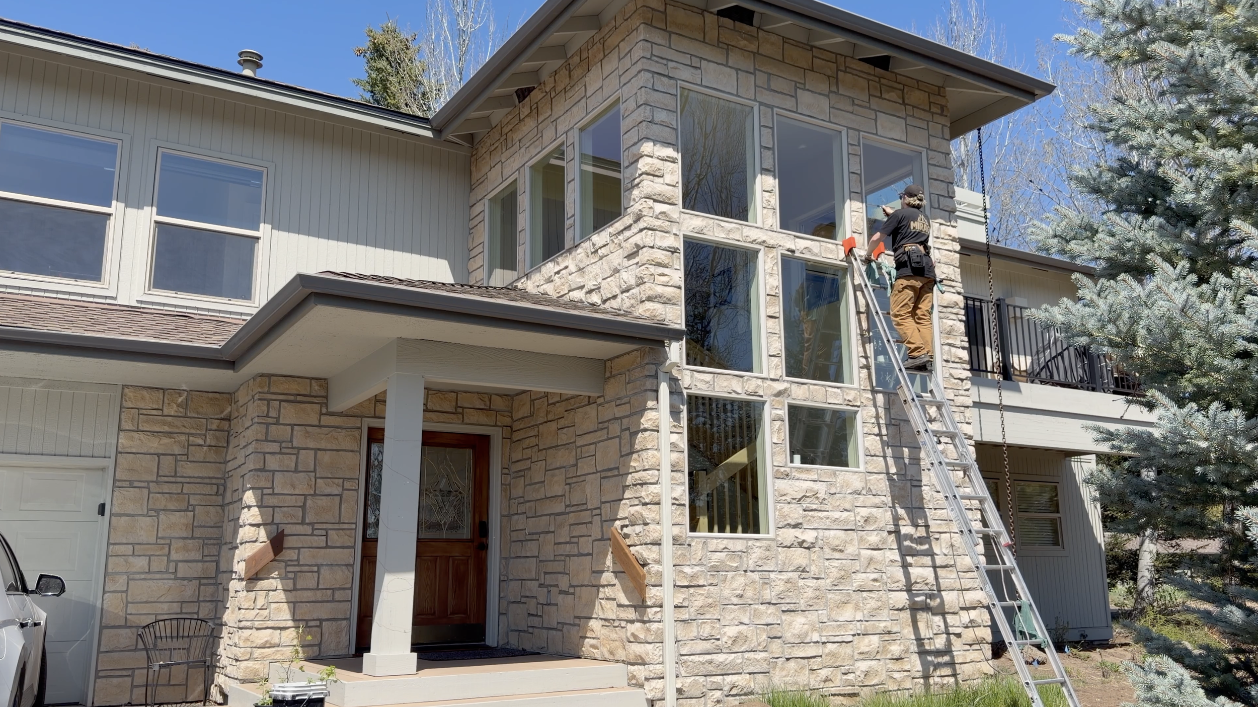 A construction worker stands on a ladder working on the exterior stone wall of a modern house, with large windows and a balcony, surrounded by trees, on a clear, sunny day.